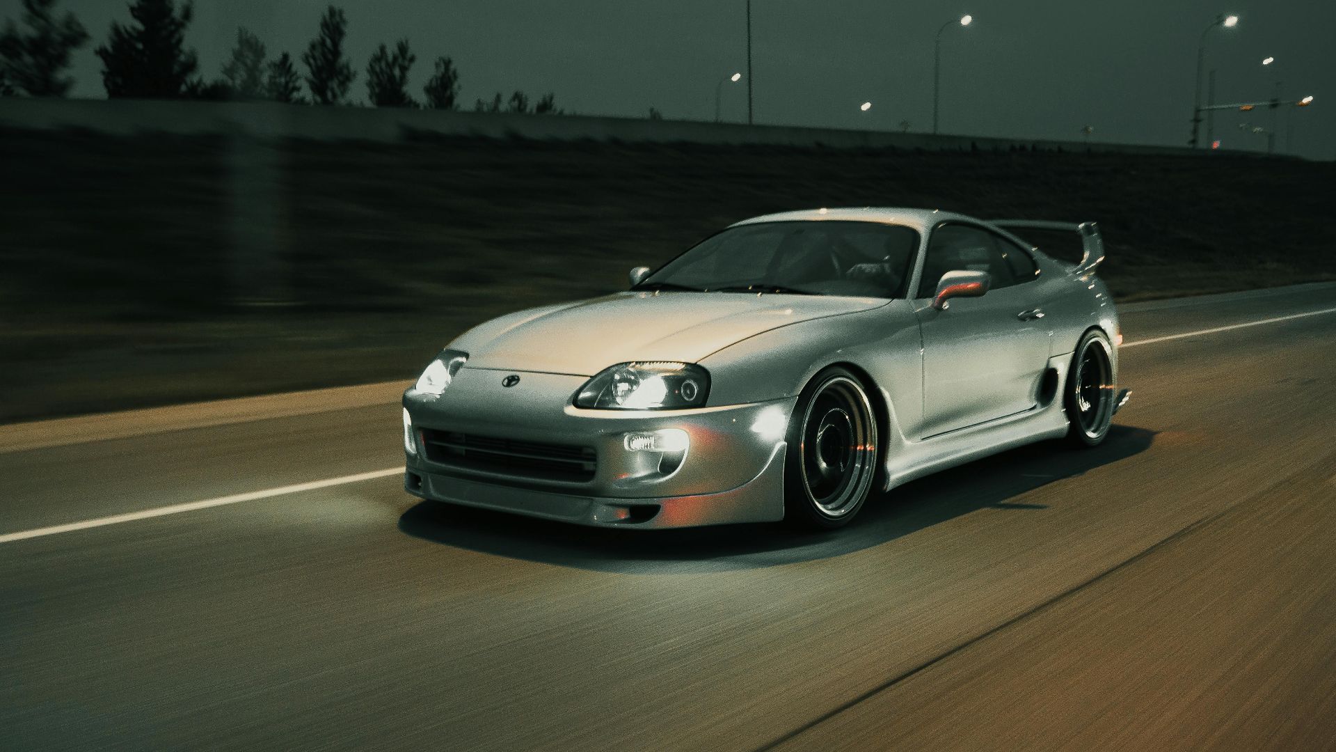 A luxurious silver sports car swiftly moves down a nighttime road, headlights illuminating the way.