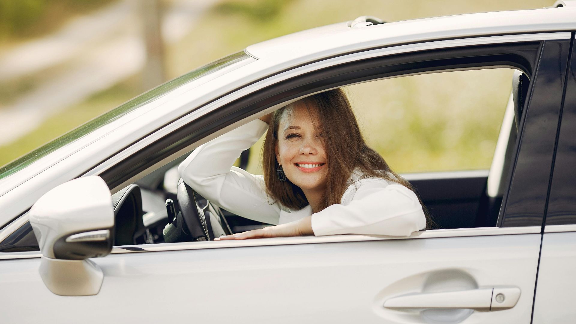 Cheerful female driver in white shirt sitting inside modern automobile and looking at camera through opened side window with smile during car trip in summer day