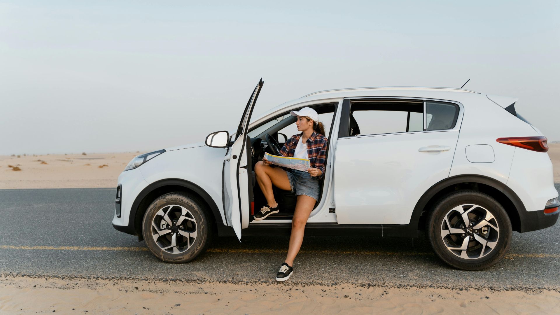 A woman sits by her SUV, exploring a map during a desert road trip under a clear sky.