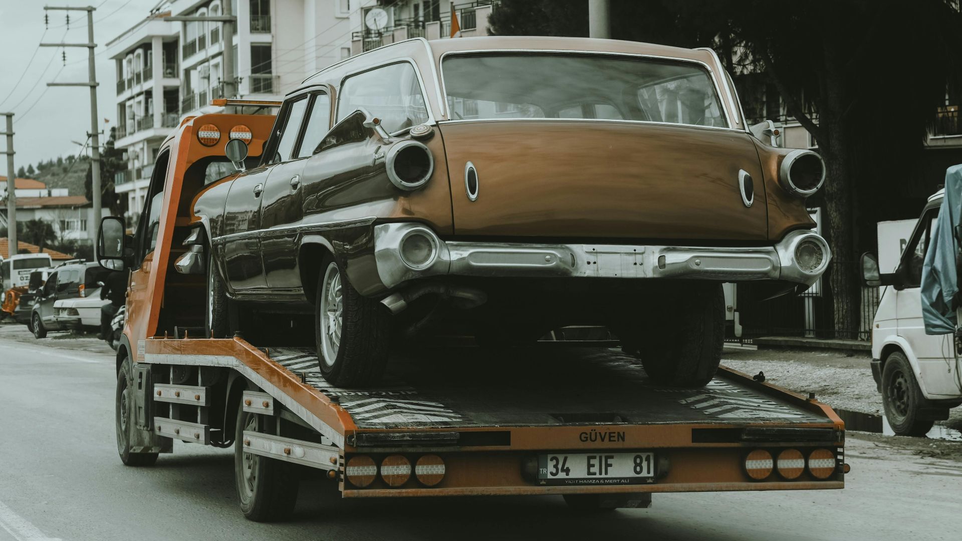 A vintage car loaded on a tow truck in a city street, surrounded by modern buildings.