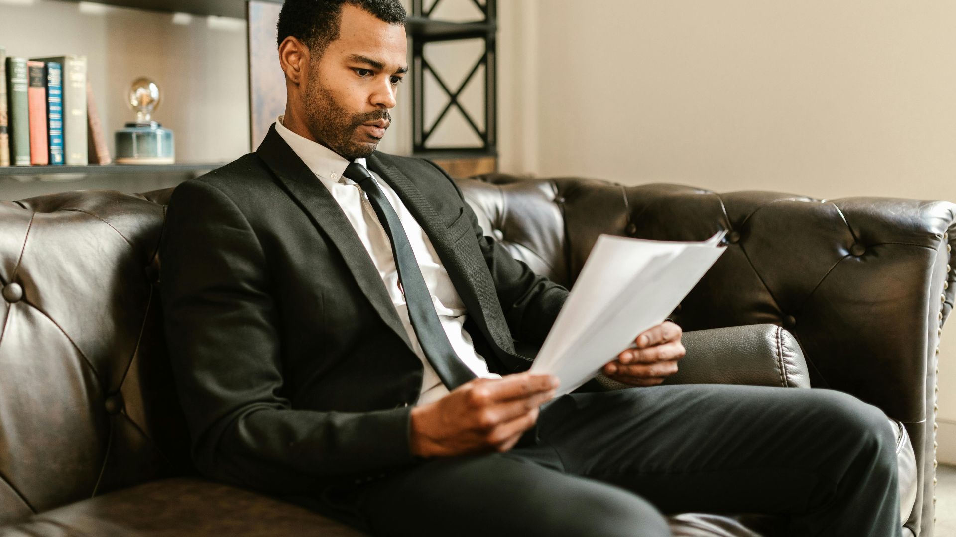 Businessman in suit reading papers on a leather sofa in a modern office setting.