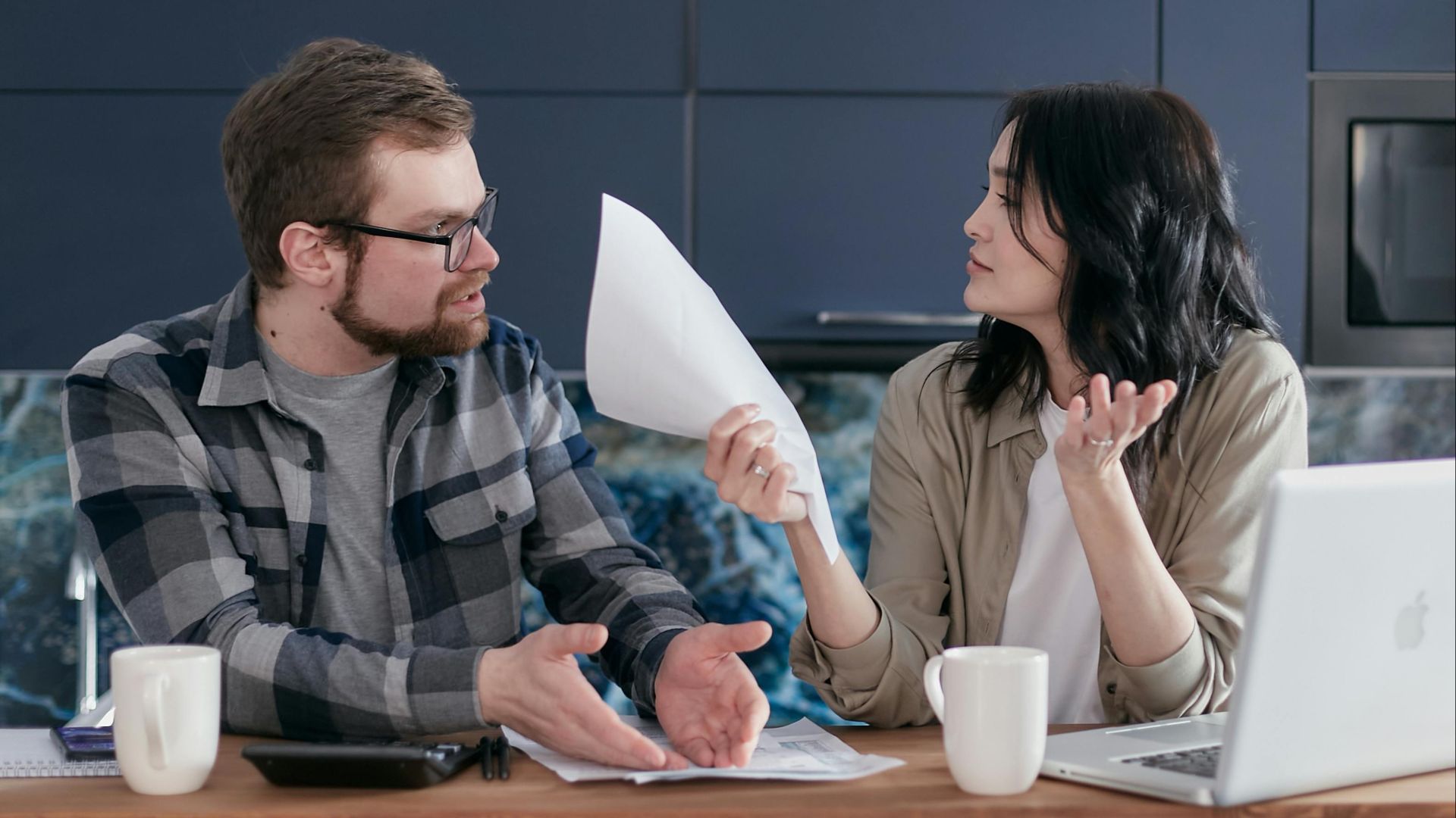 A couple sits at a table, discussing finances with papers and a laptop, looking concerned.