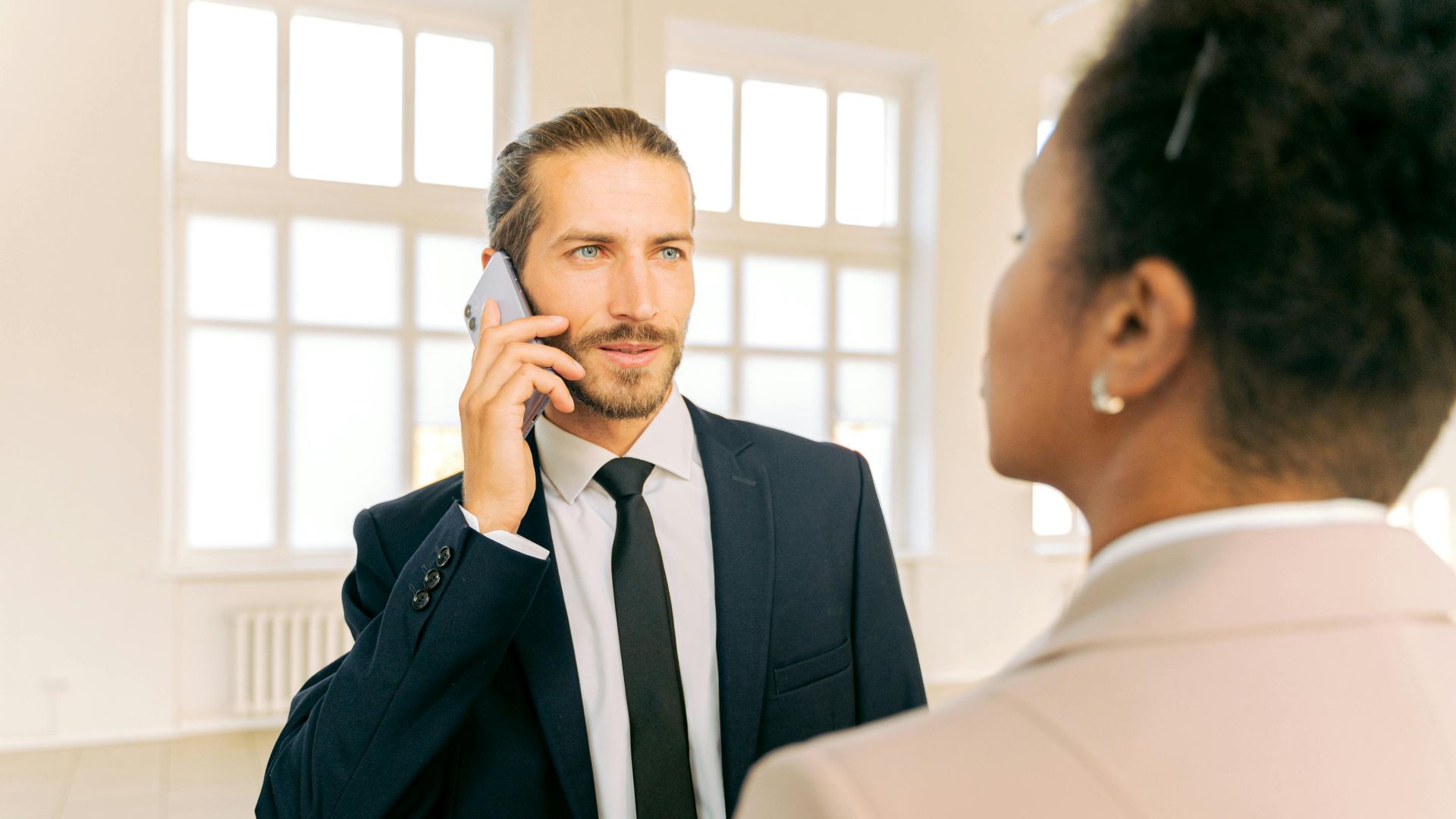 Man in business suit talking on phone with colleague in bright office setting.