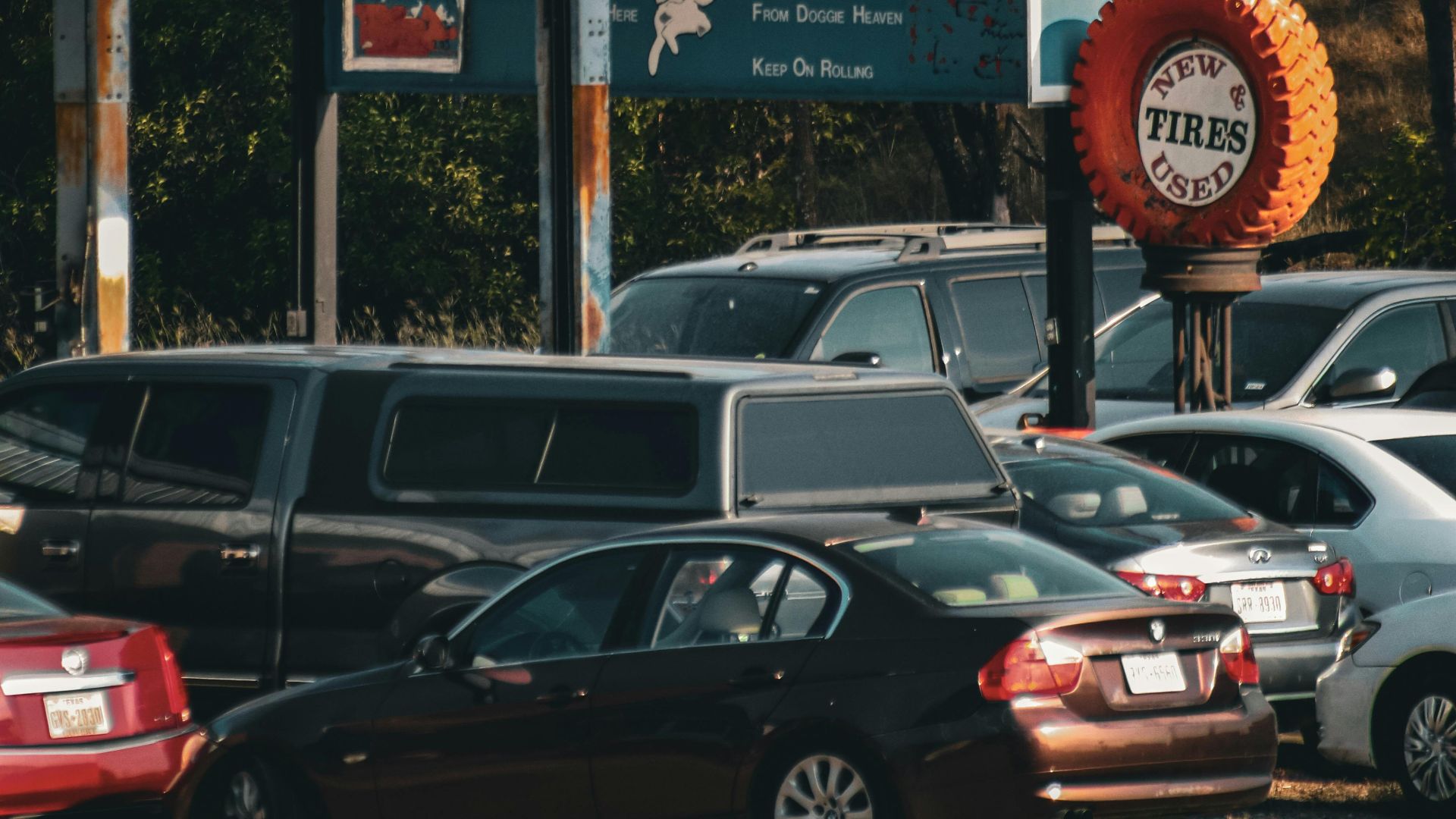 A parked car lot with a used tire shop surrounded by lush trees in autumn.