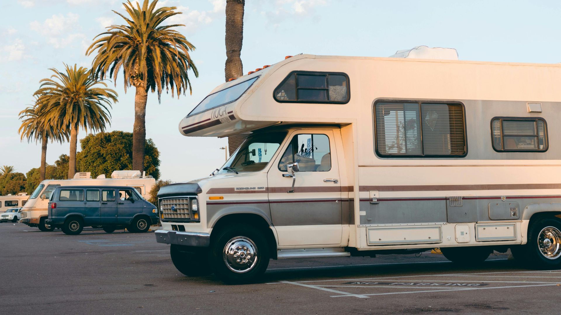 RVs parked under palm trees in a bright daylight urban parking lot setting.