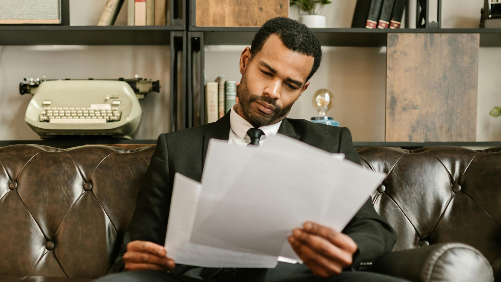 Adult man in a suit reading paperwork on a leather couch in a stylish office setting.