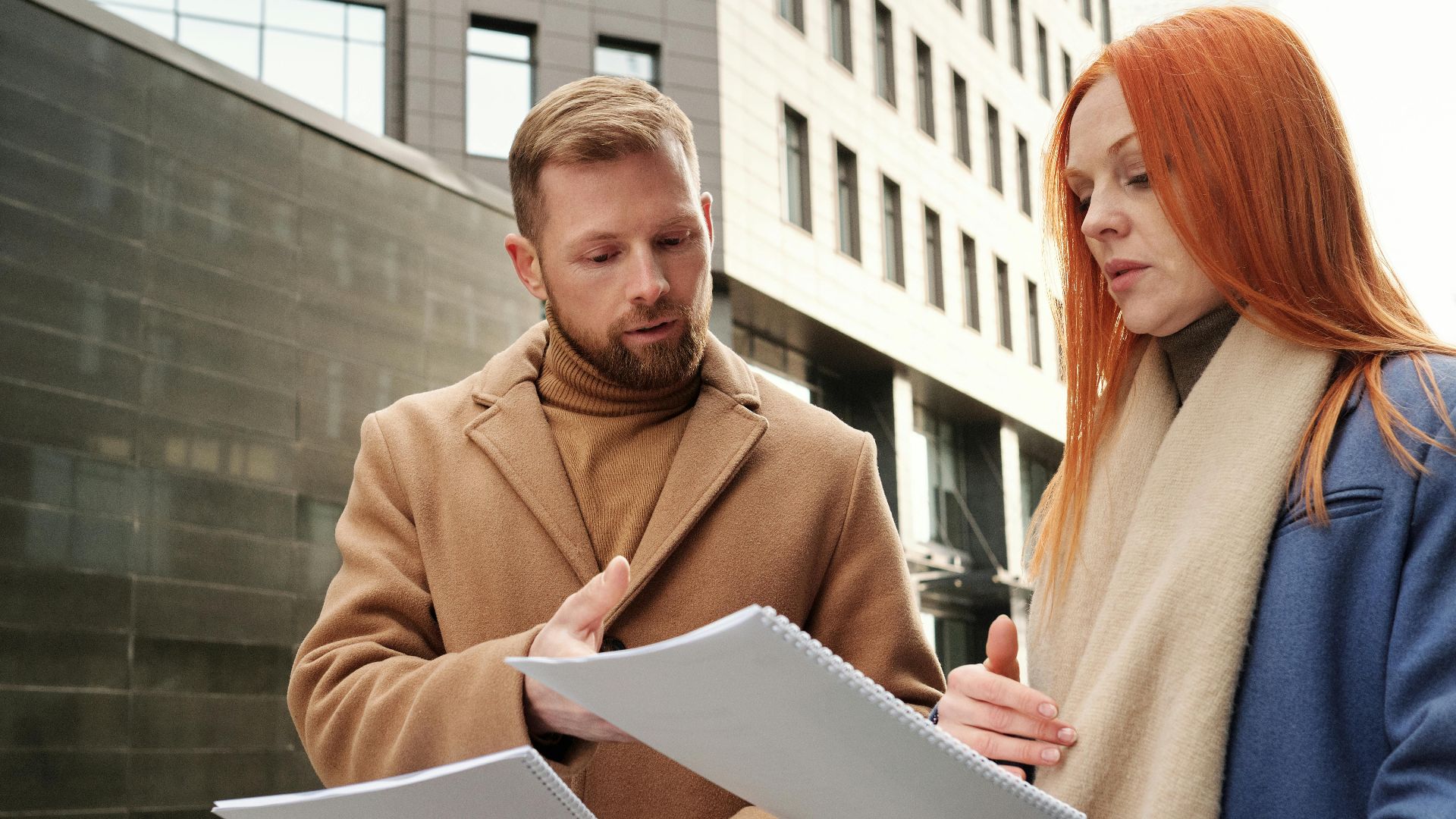 Two business professionals in coats discuss documents in an urban setting.