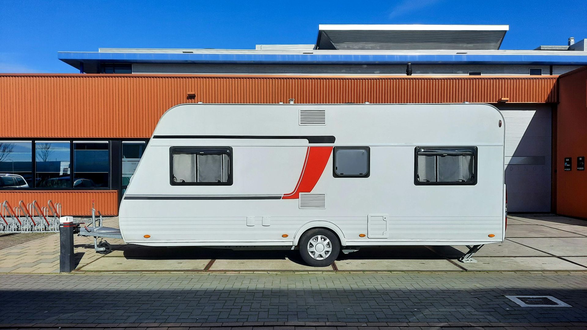 A sleek white caravan with red accents parked next to an industrial building under a clear blue sky.