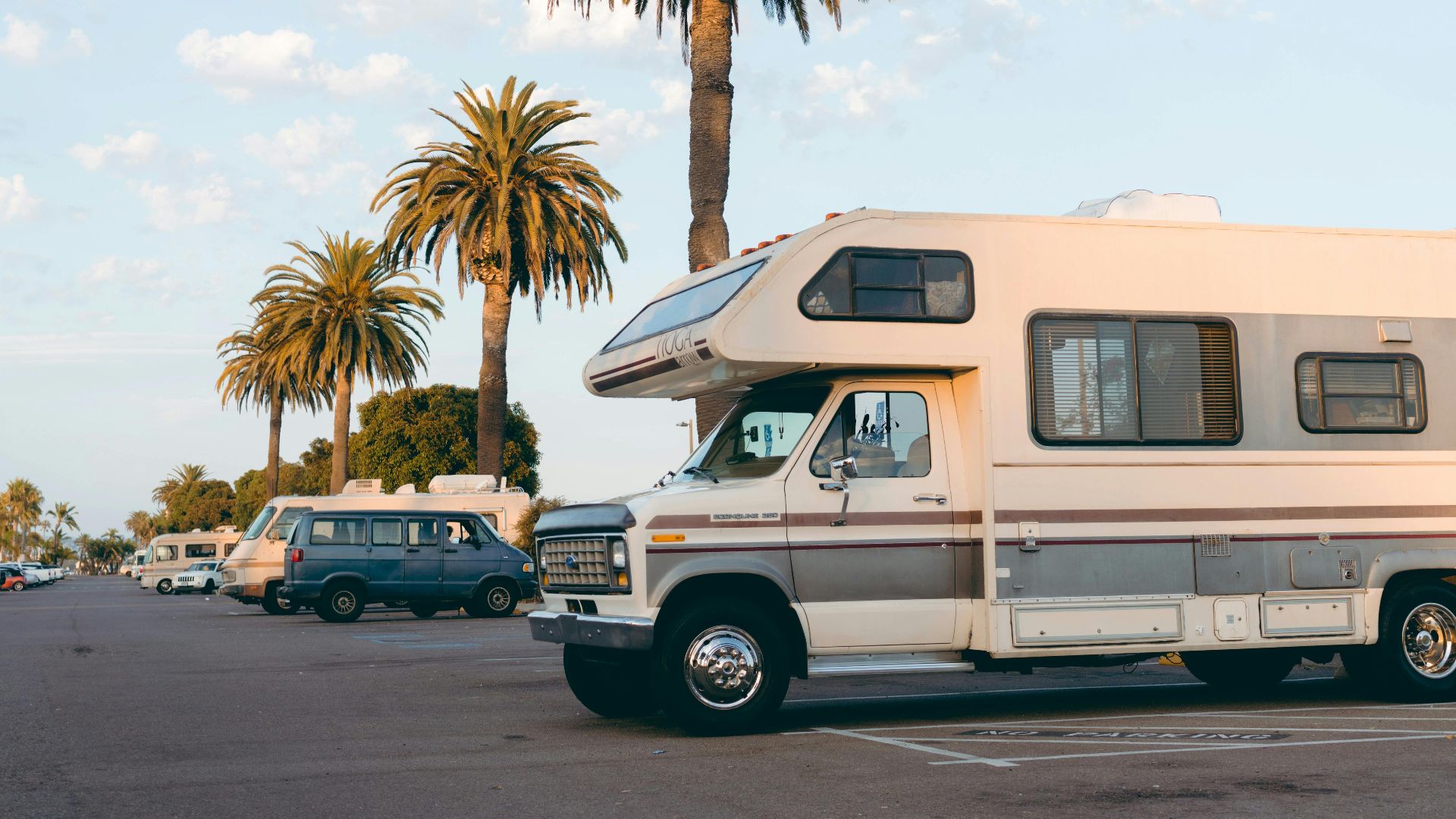 RVs parked under palm trees in a bright daylight urban parking lot setting.