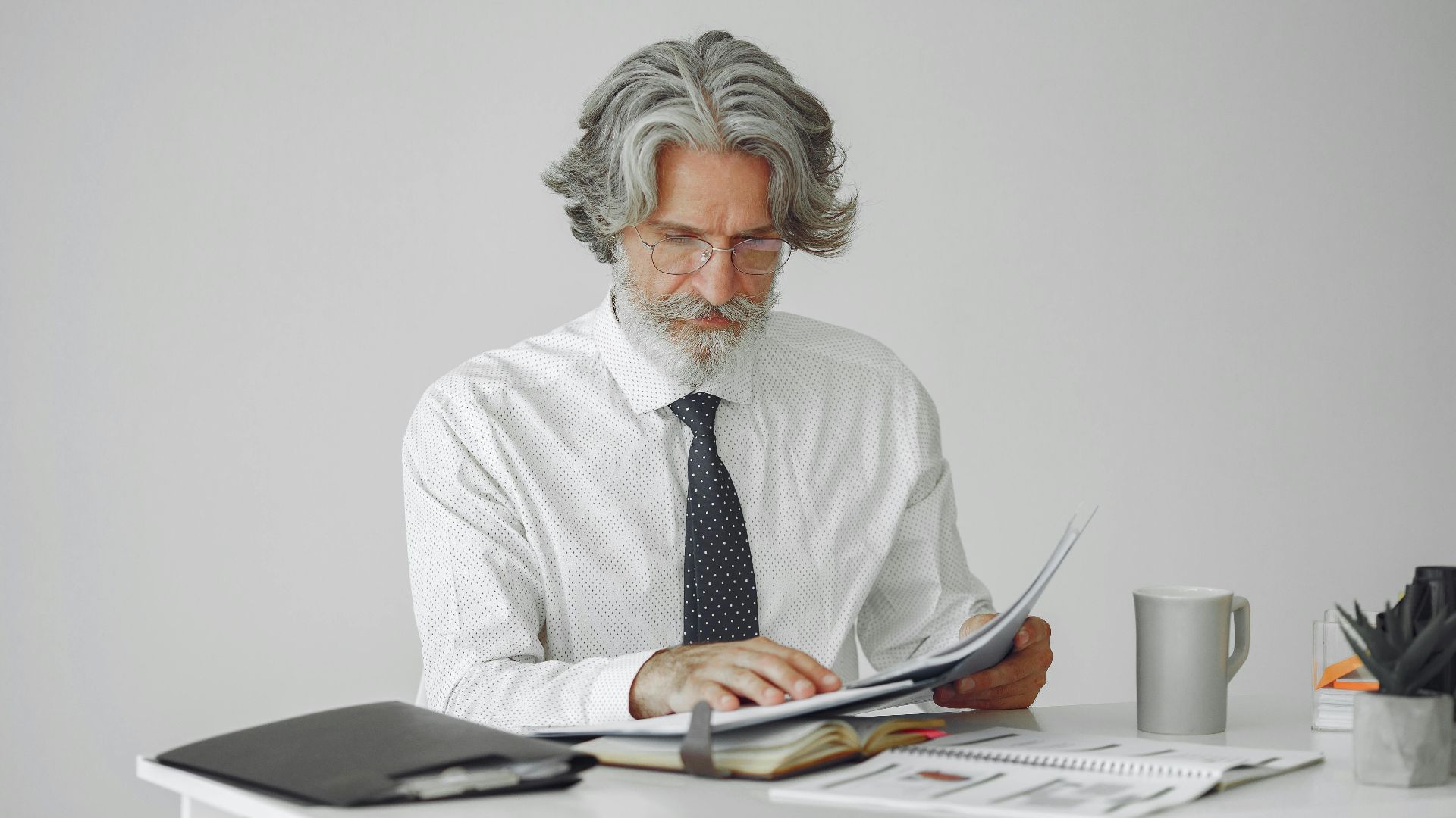 An elderly businessman reviewing documents at his desk in a modern office setting.