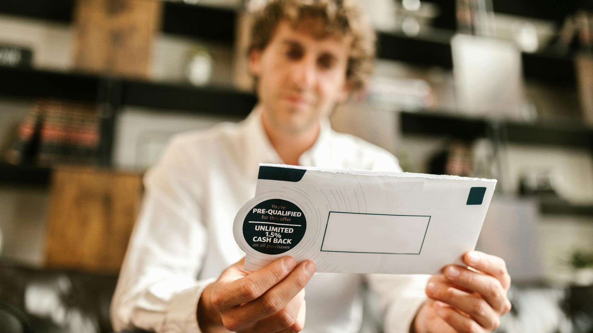 A man sitting indoors, reading an envelope with a cash back offer, blurred background.