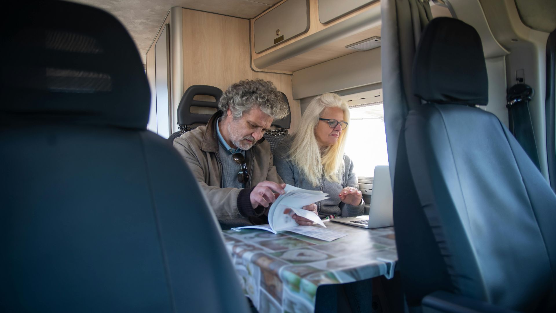 An adult couple working indoors in a caravan, using a laptop and reviewing documents.
