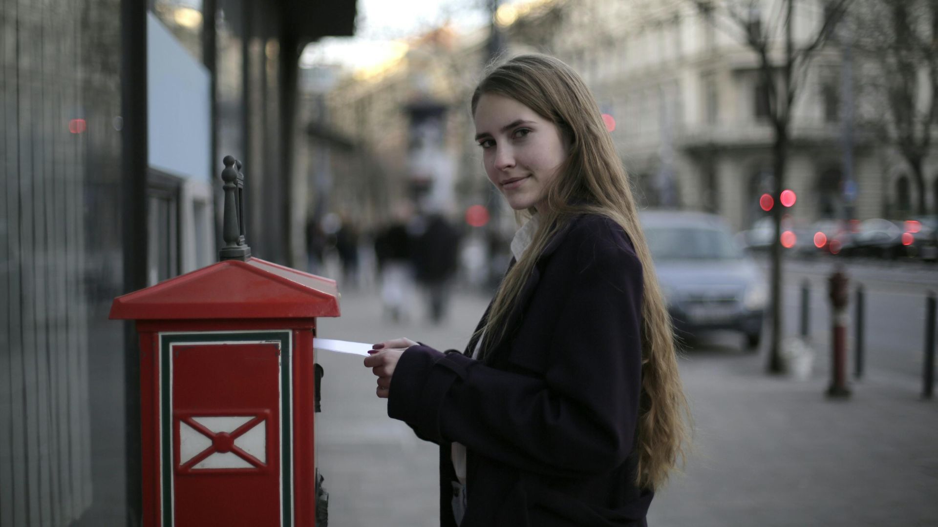 Young woman mailing a letter in a red mailbox on a city street during the day.