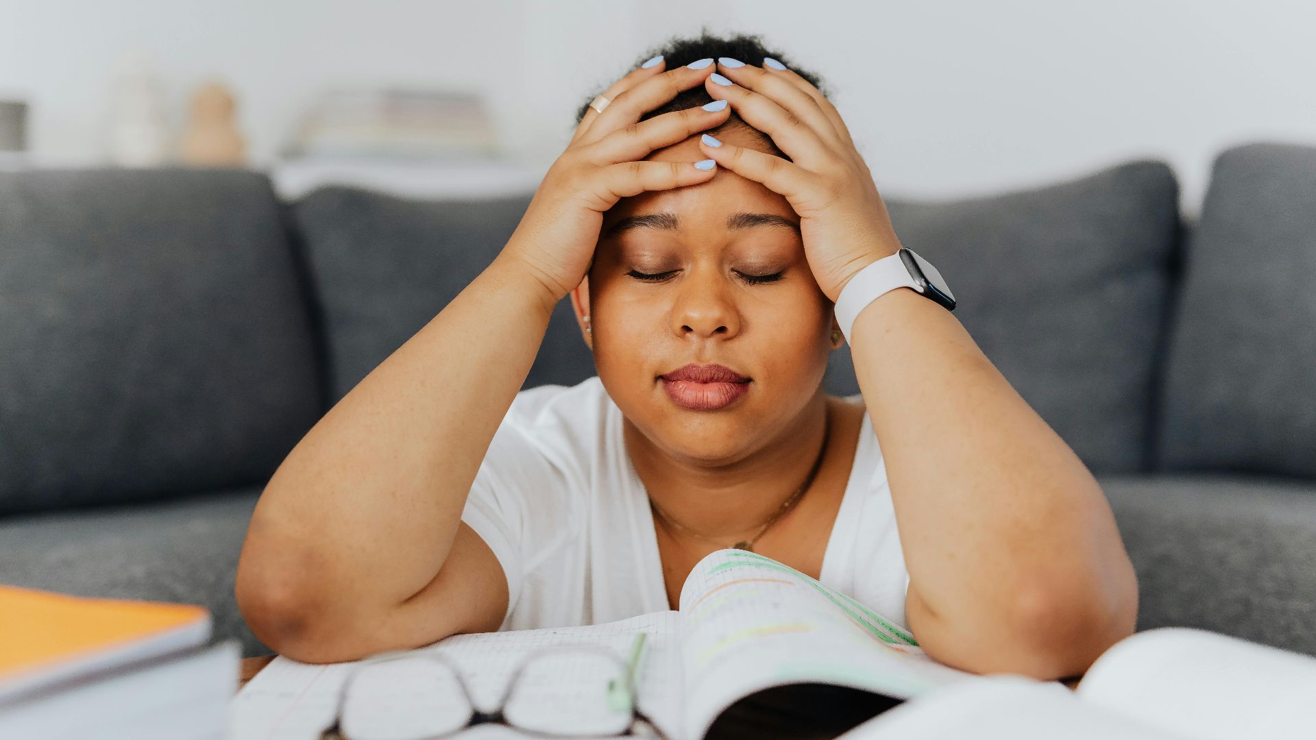 A woman with eyes closed holds her head in stress, surrounded by books in an indoor setting.