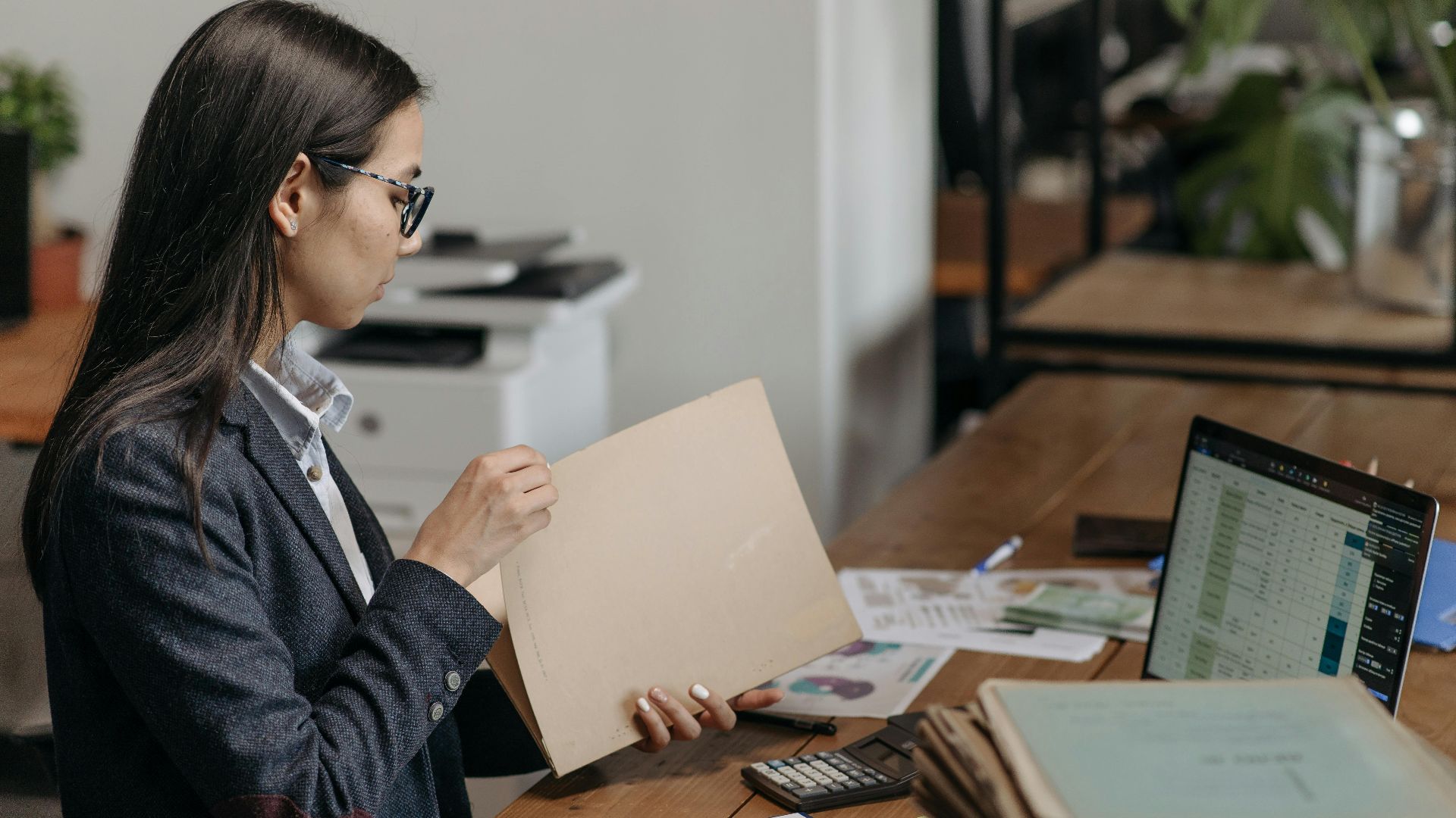 A professional woman reviewing documents at her office desk with a laptop and files.