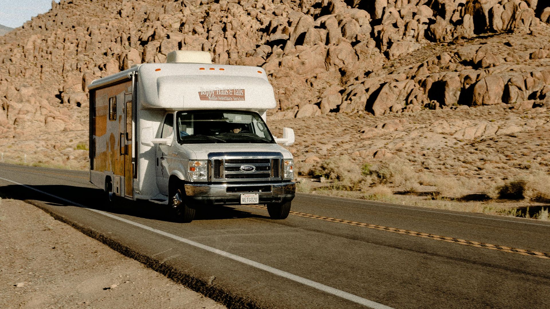 RV driving through a scenic desert highway with rocky mountains under a clear sky.