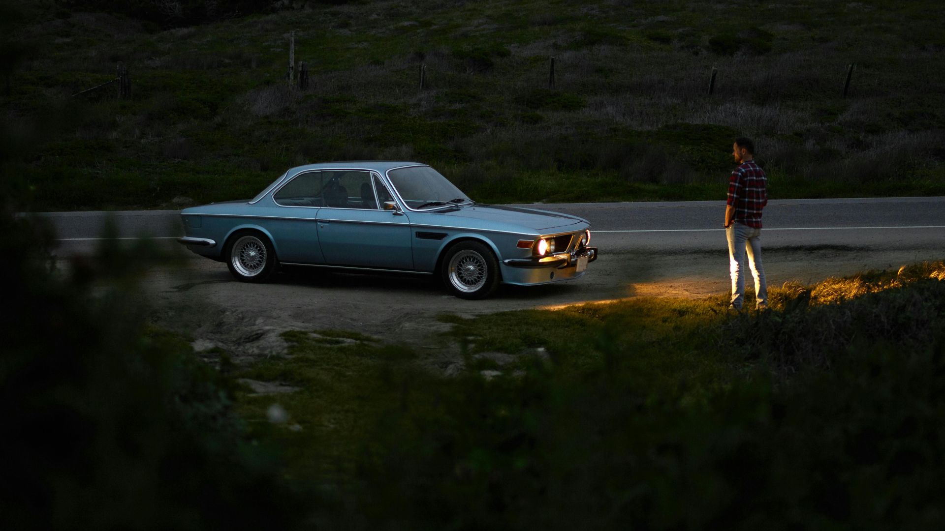 Back view of full length anonymous man in casual outfit standing on roadside in front of parked retro car with lights on near field in twilight
