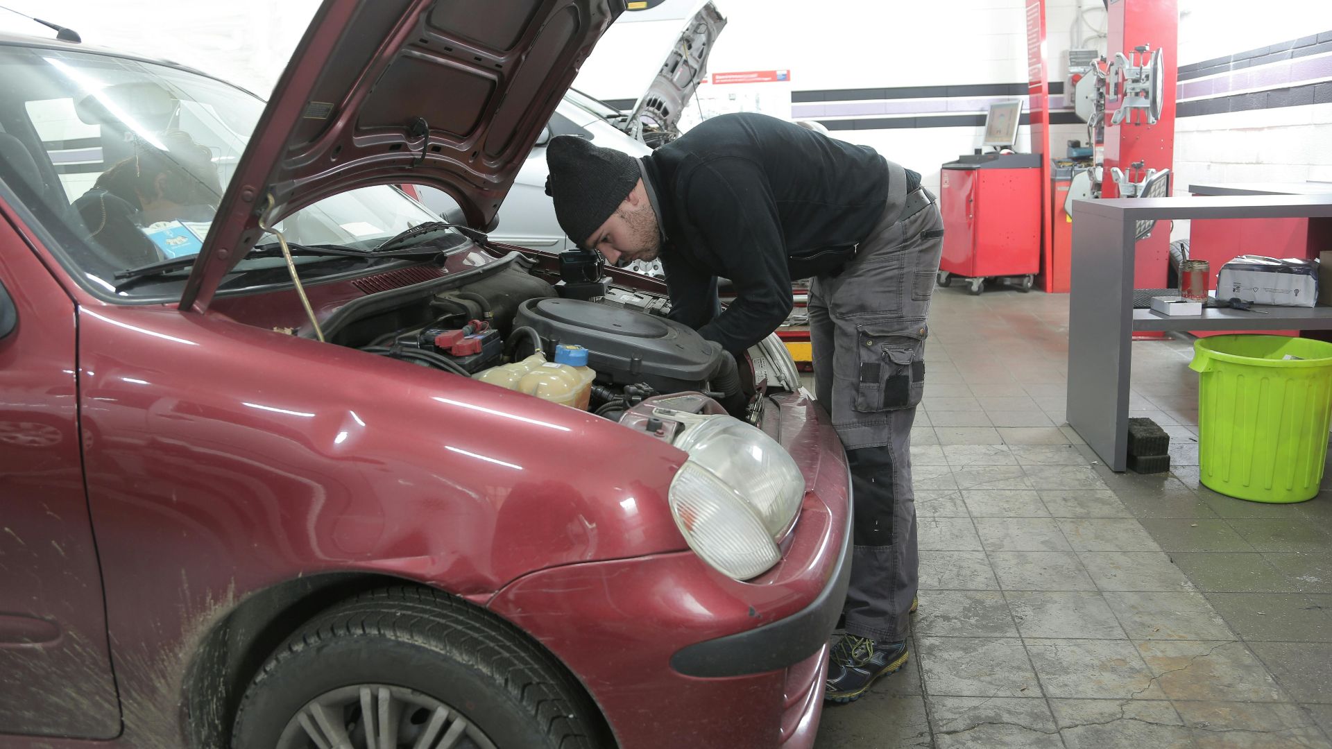 Mechanic performs engine inspection on a red car indoors, in a garage workshop.