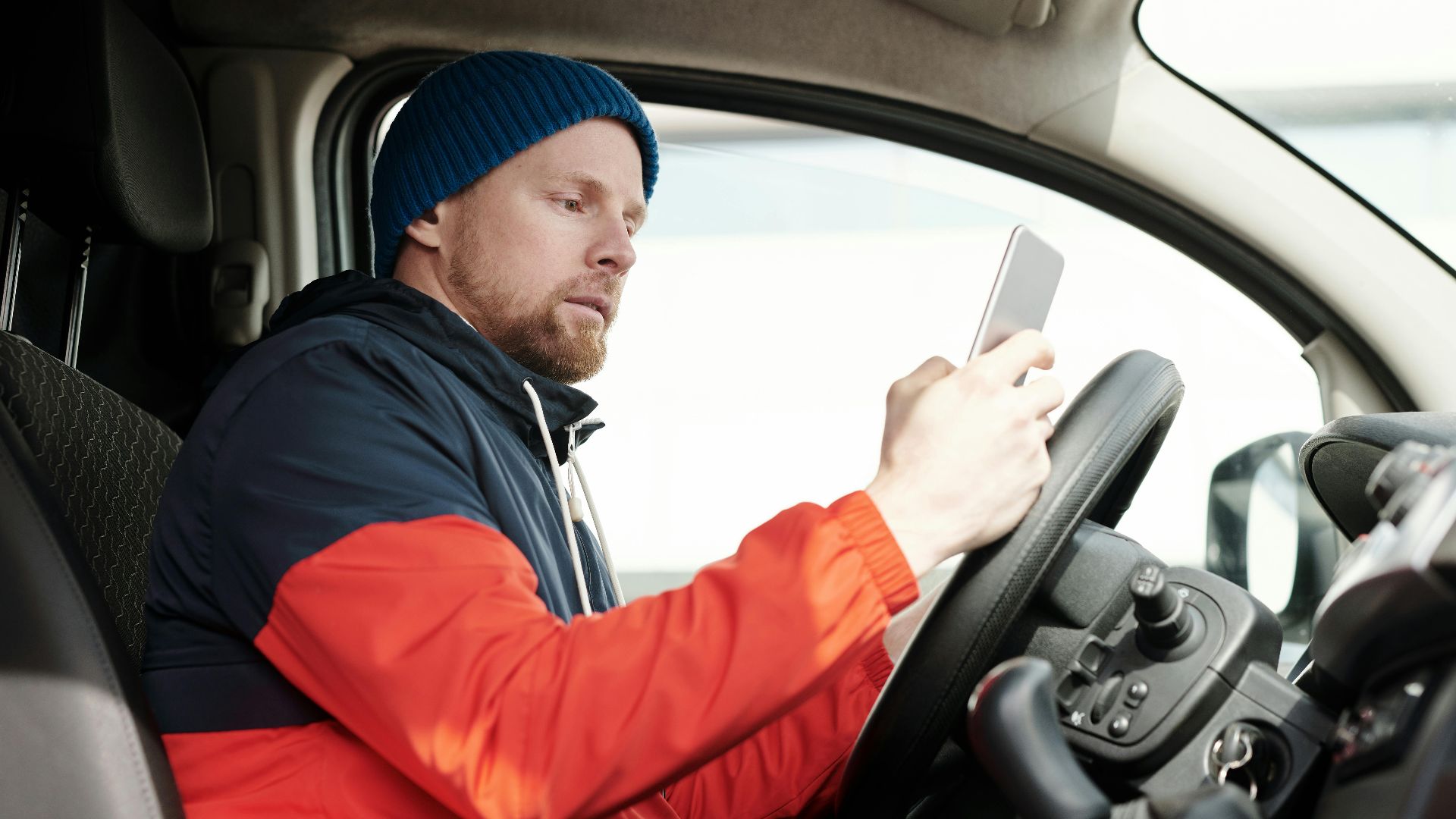 Man in a parked car using a smartphone, wearing a blue beanie and red jacket. Daytime driving scene.