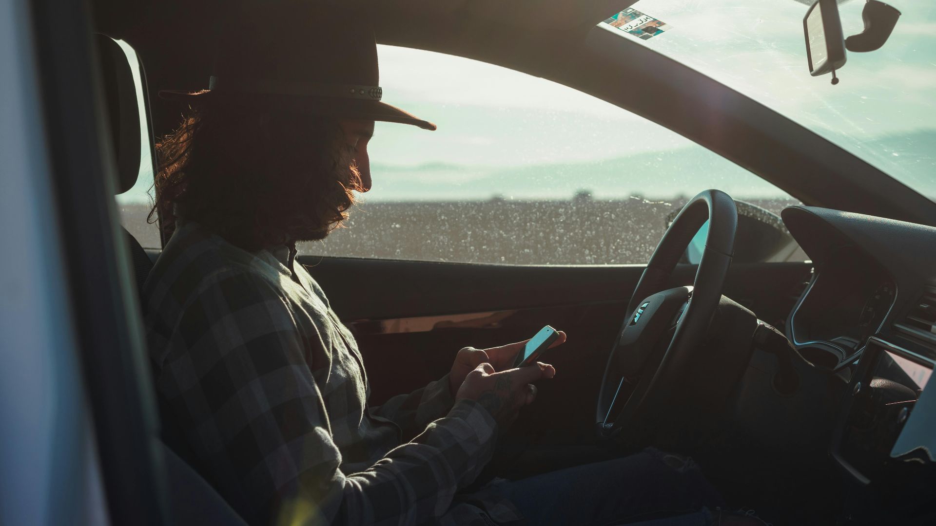 Profile of a man with a hat using a phone inside a car during the day.