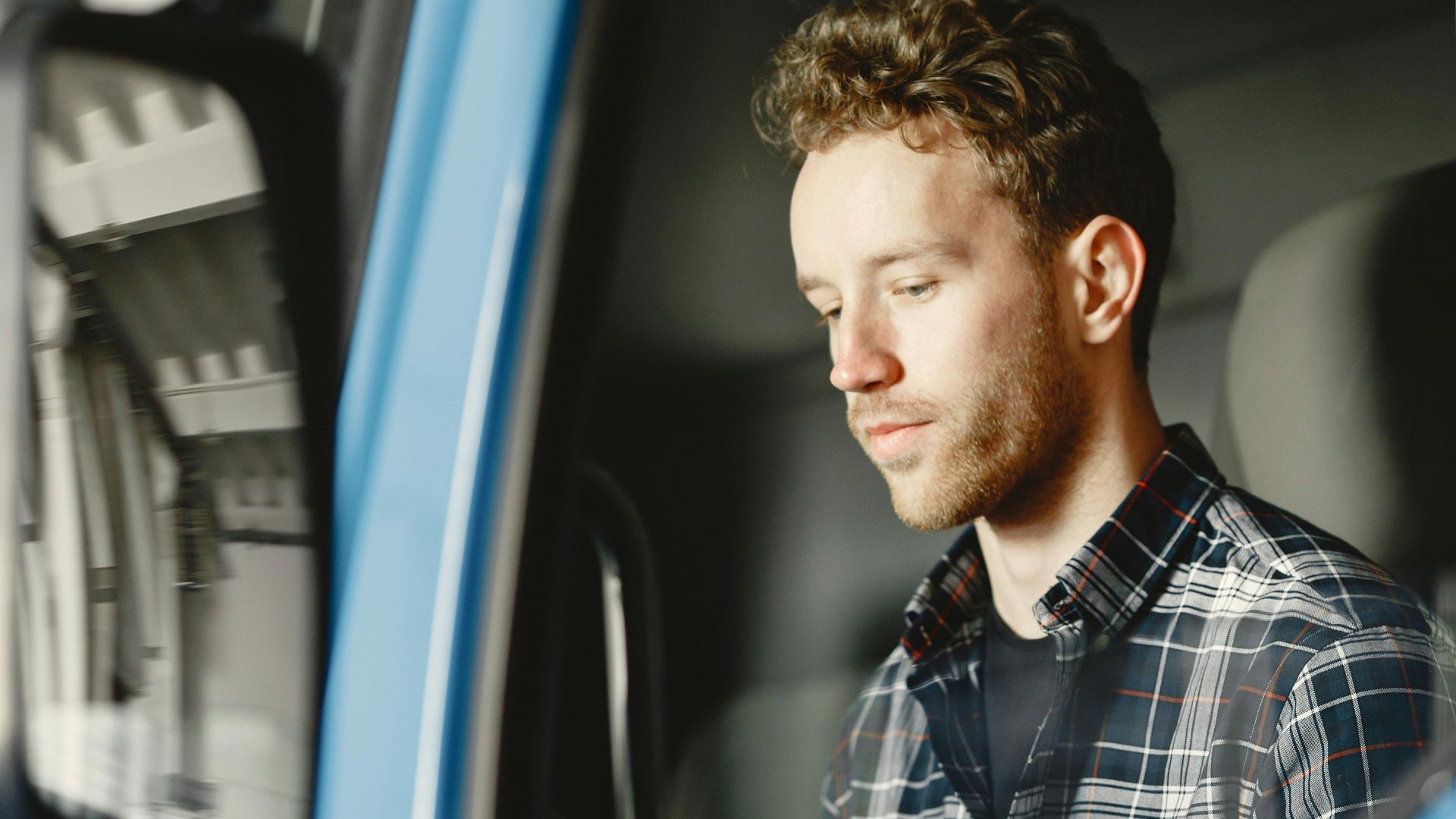 A young man in a checked shirt sitting inside a blue truck's cabin.
