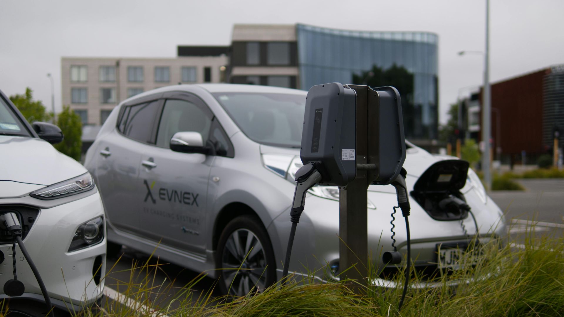 Electric vehicles at a charging station in Christchurch parking lot with modern architecture.