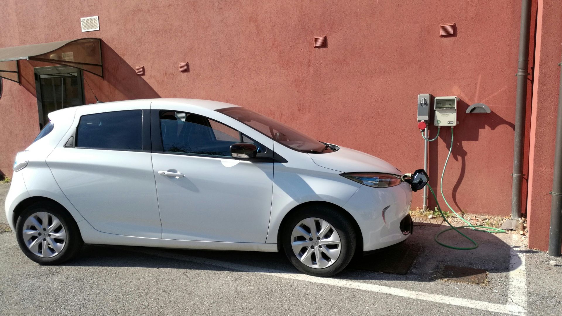White electric vehicle charging at station against a red wall in San Bartolomeo di Breda di Piave, Italy.