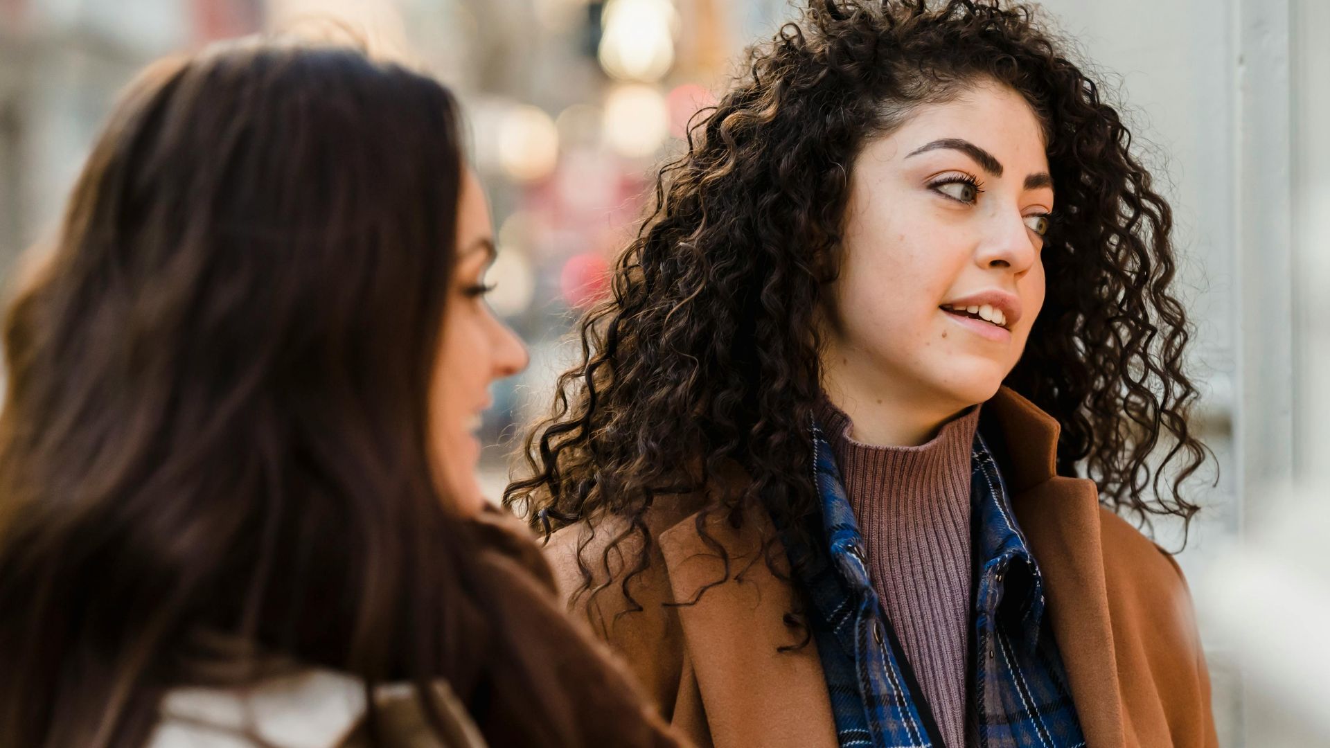 Optimistic young multiracial girlfriends with long dark hairs in warm clothes smiling while standing on street during weekend in city