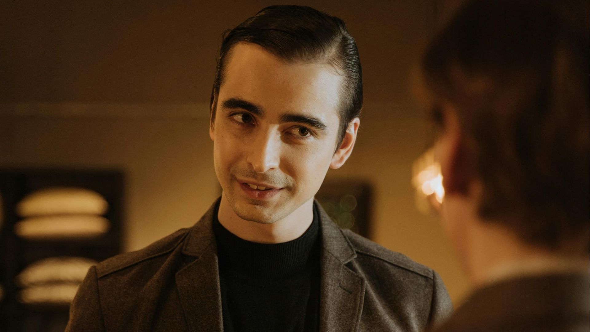 Young adult man in a suit conversing in a warmly lit indoor setting.