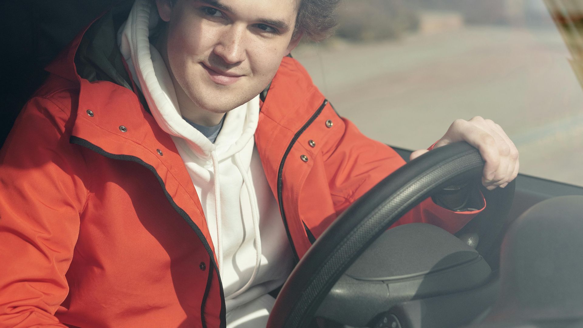 A young adult man smiling while sitting behind the steering wheel of a vehicle.