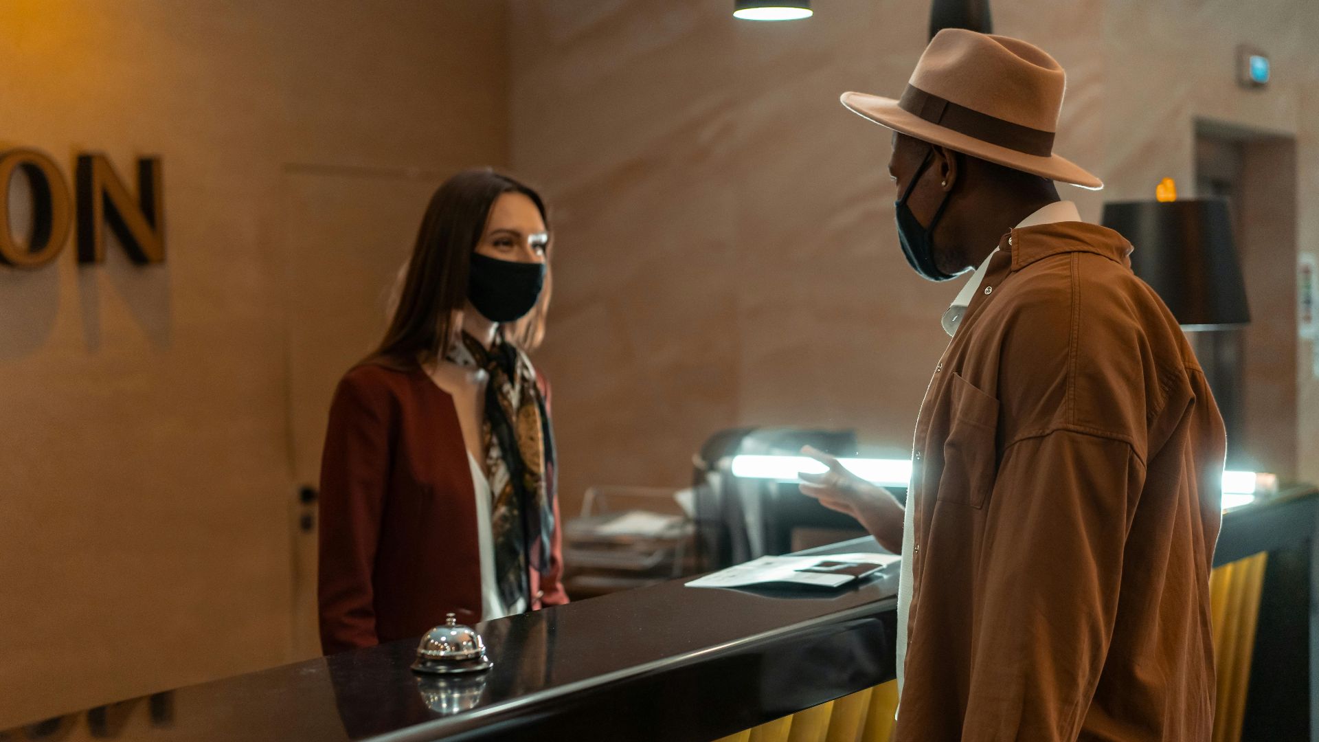 A man at a hotel reception talking to a staff member. Both wear face masks.