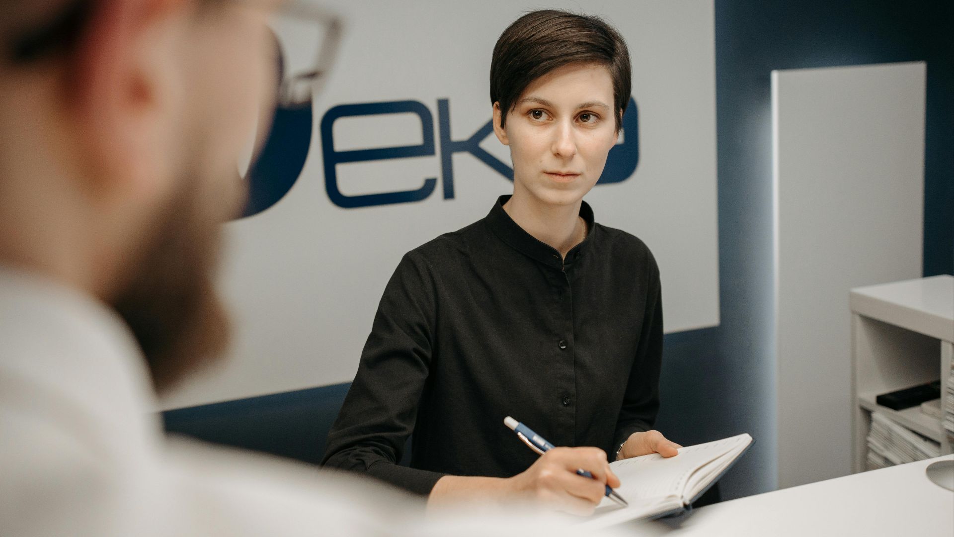 A receptionist at a clinic desk writing notes during a consultation.