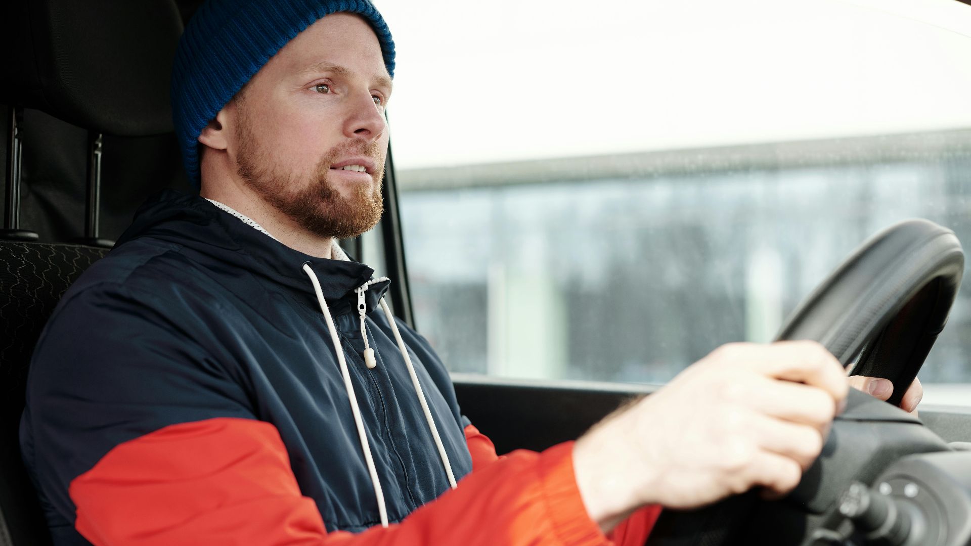 A bearded man wearing a hoodie and knitted cap driving a car, focused and attentive.