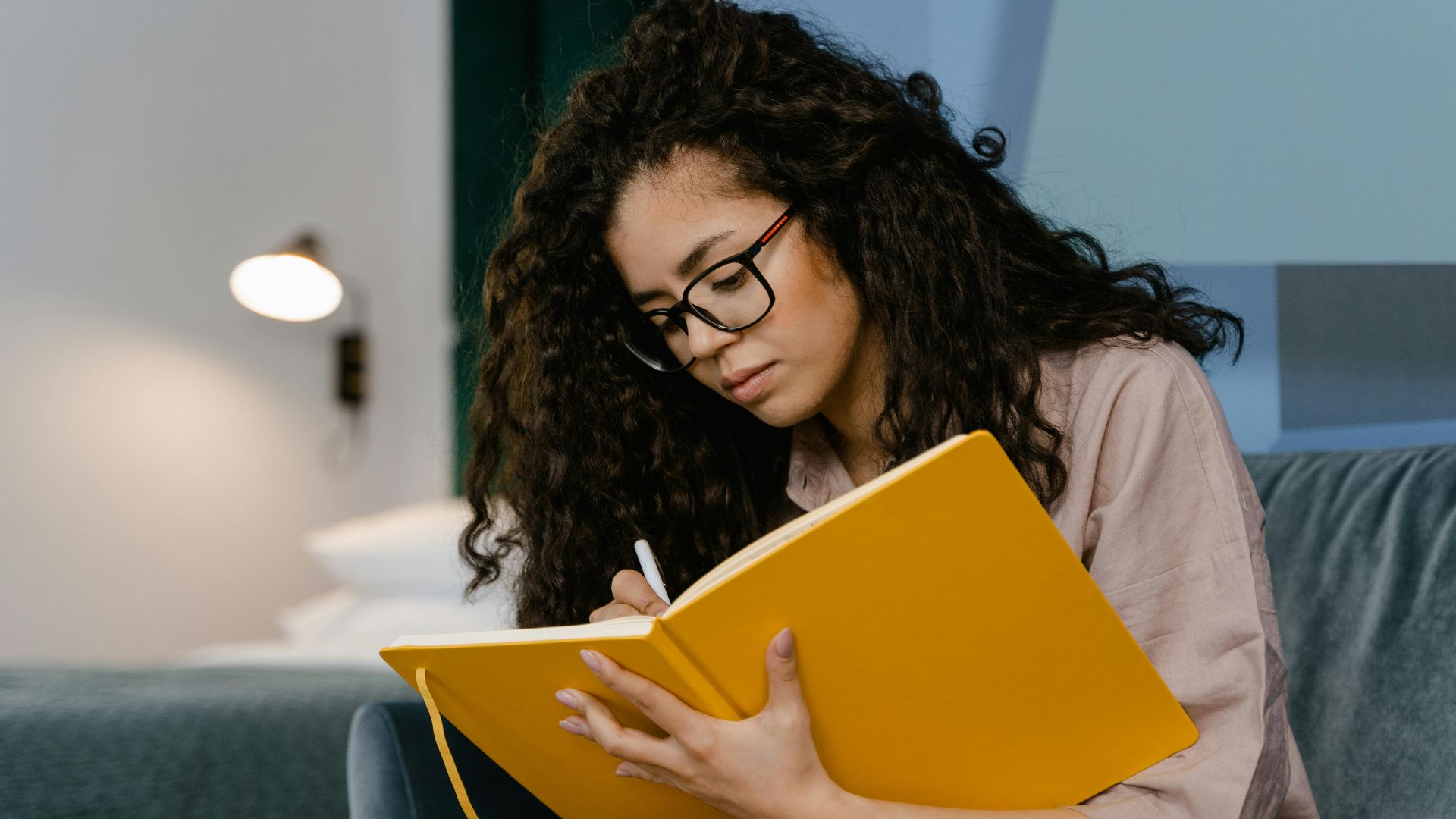 Young woman with curly hair writes in a yellow notebook, seated indoors.