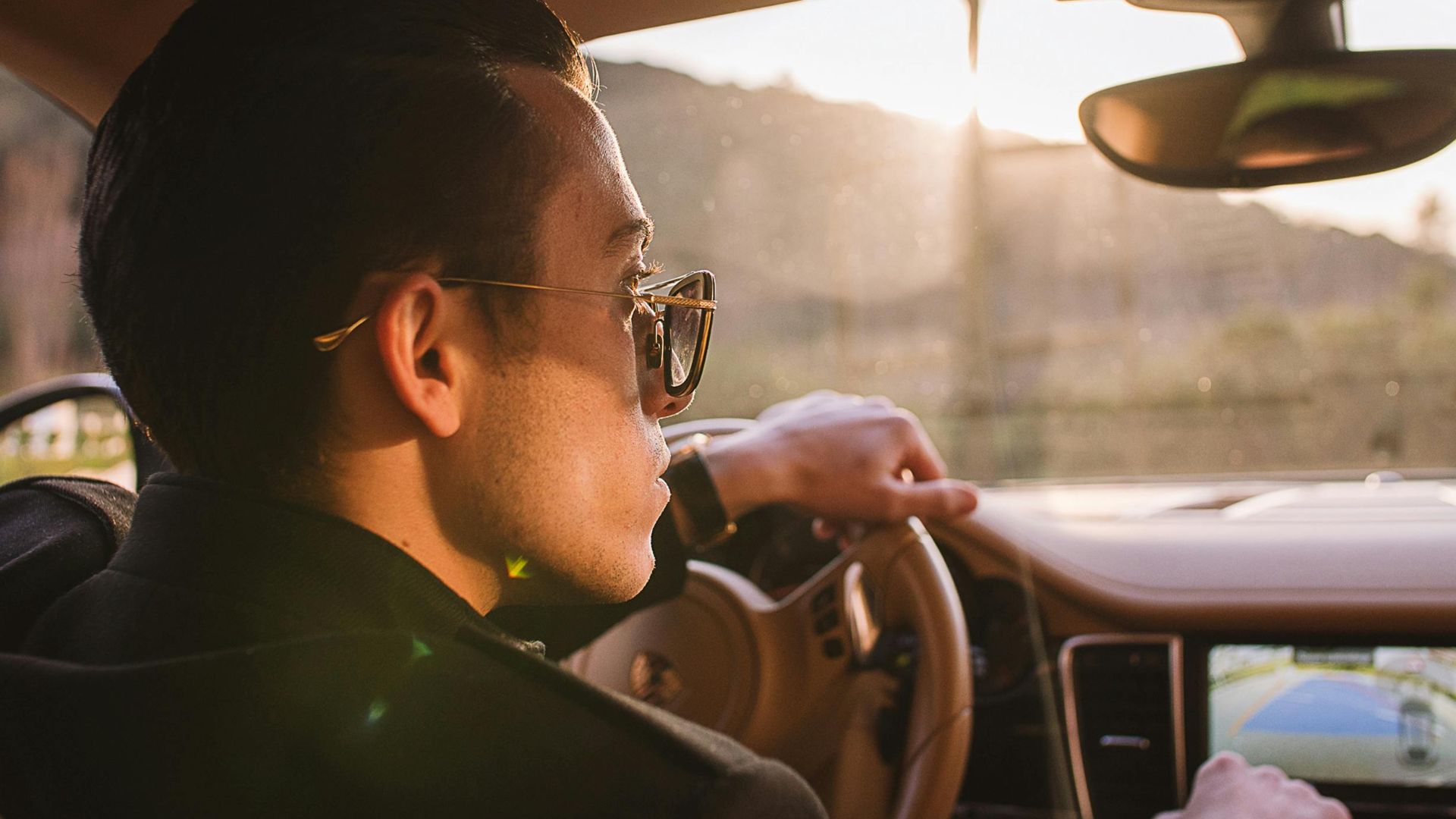 Man driving luxury car at sunset wearing sunglasses, showcasing elegant car interior and confident style.
