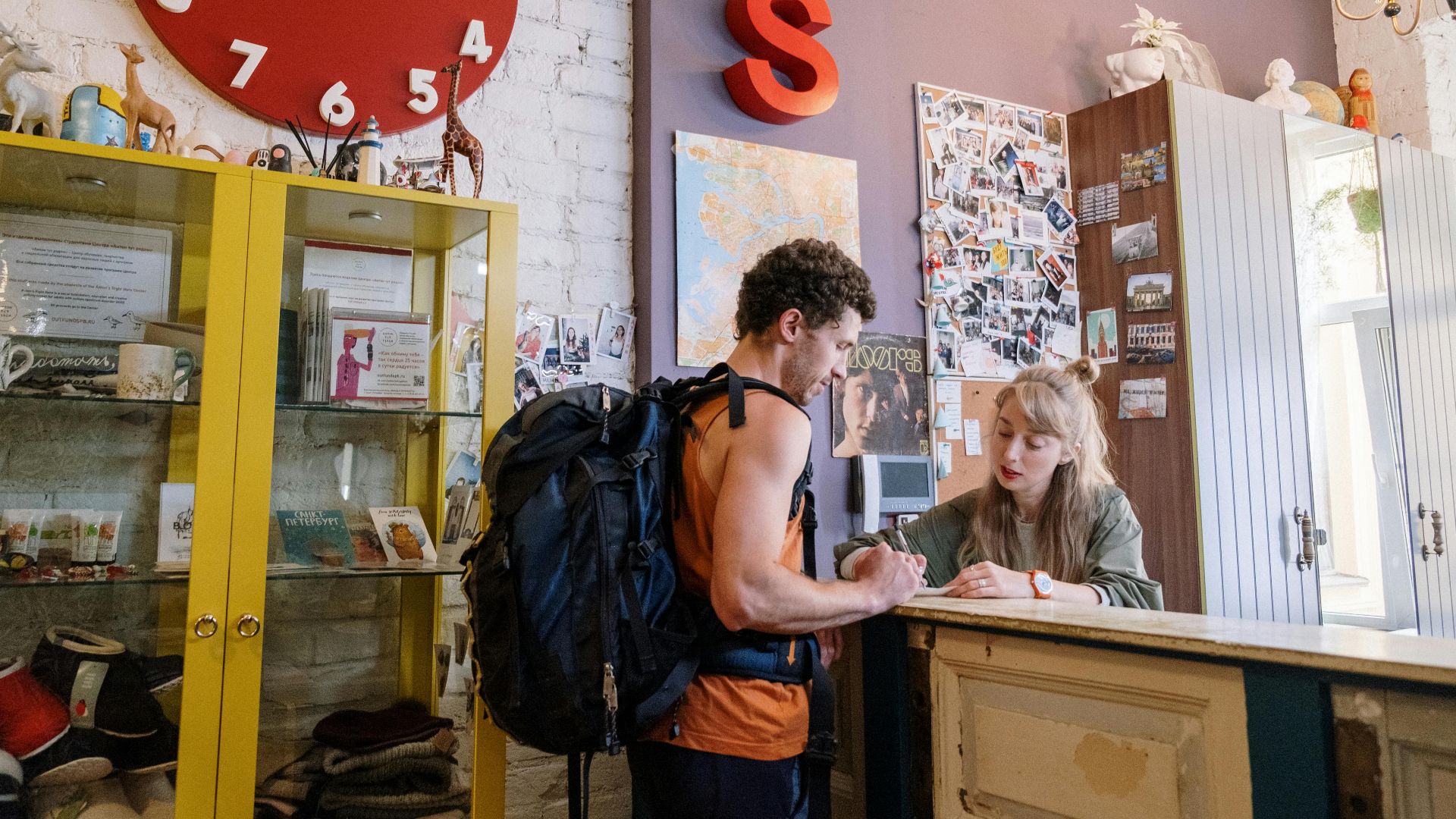 A backpacker at a hostel front desk getting assistance from a receptionist.