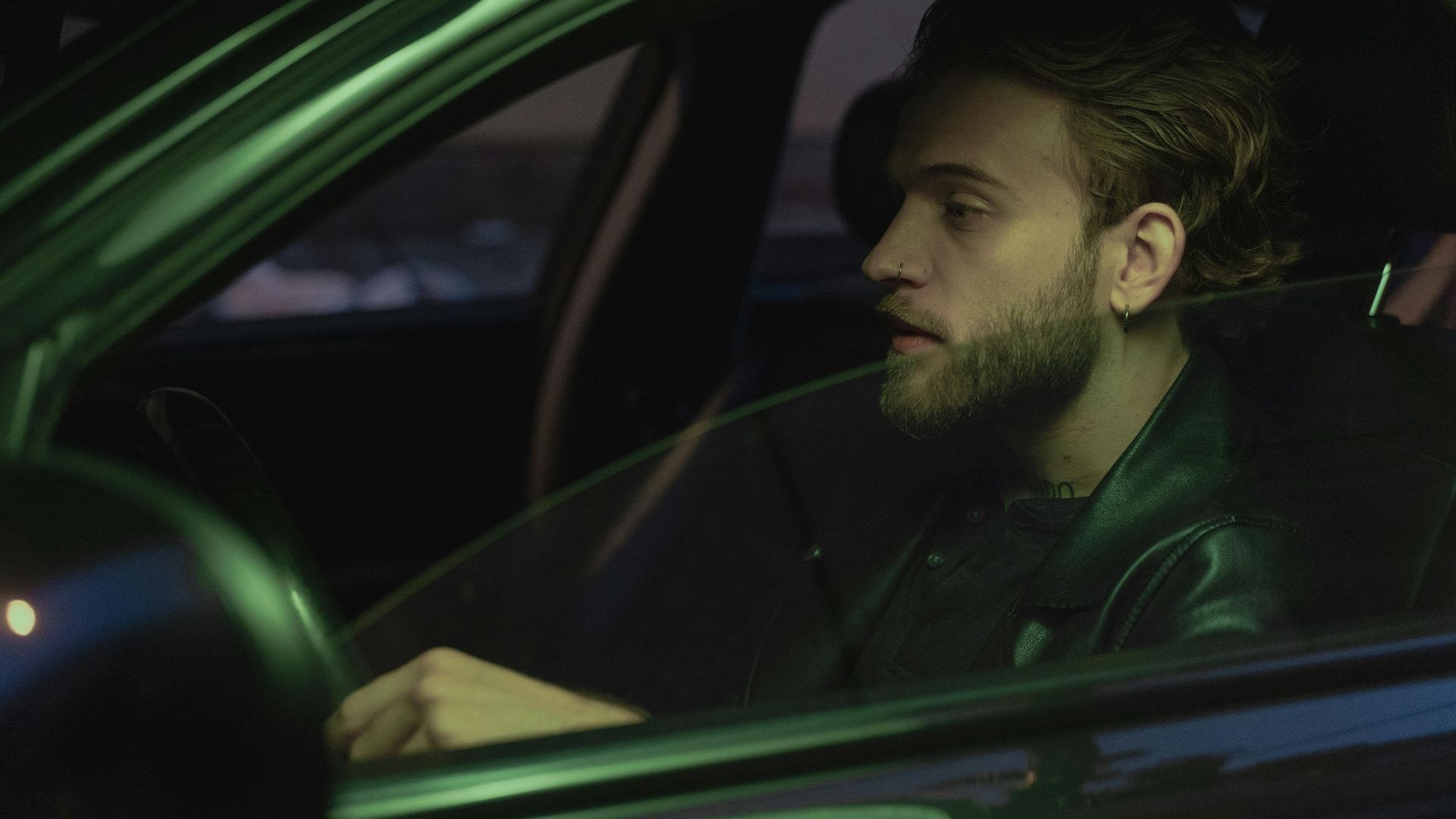 A young man with a beard drives at night, wearing a leather jacket.