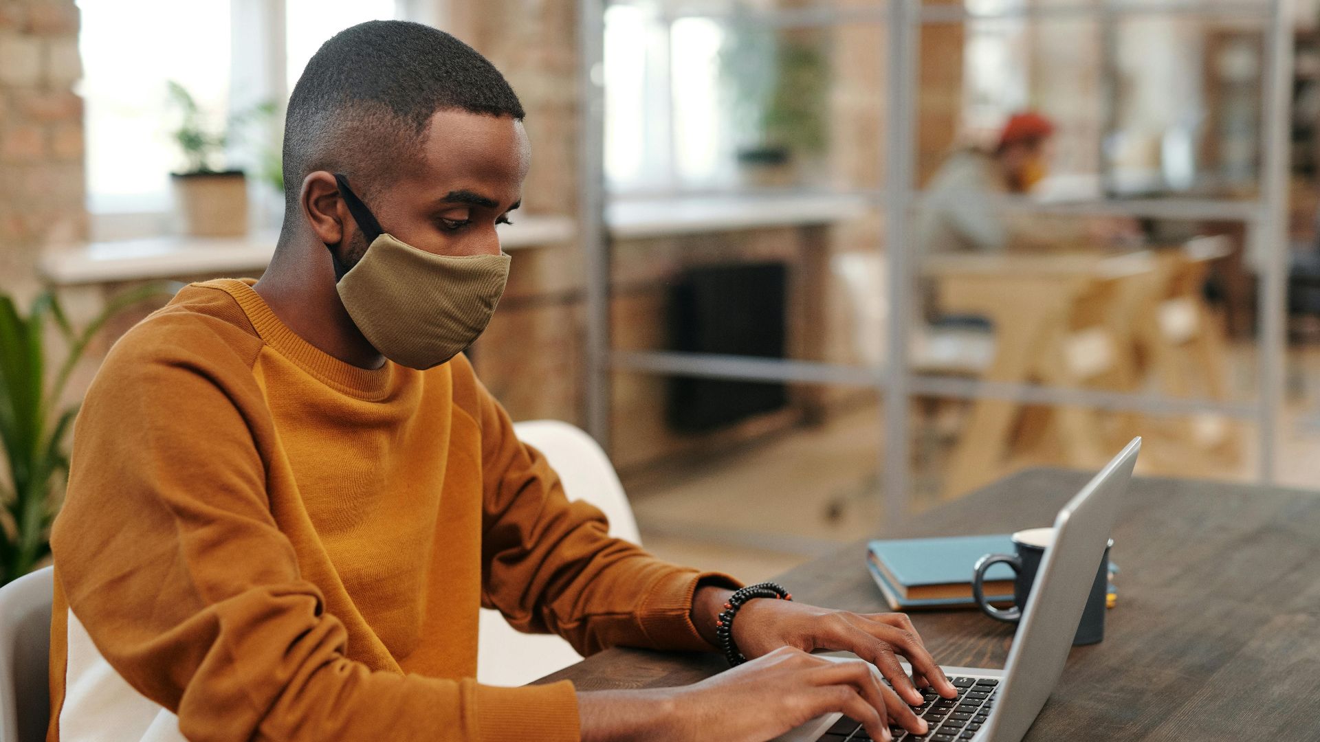 Man wearing a facemask typing on a laptop indoors with blurred background.