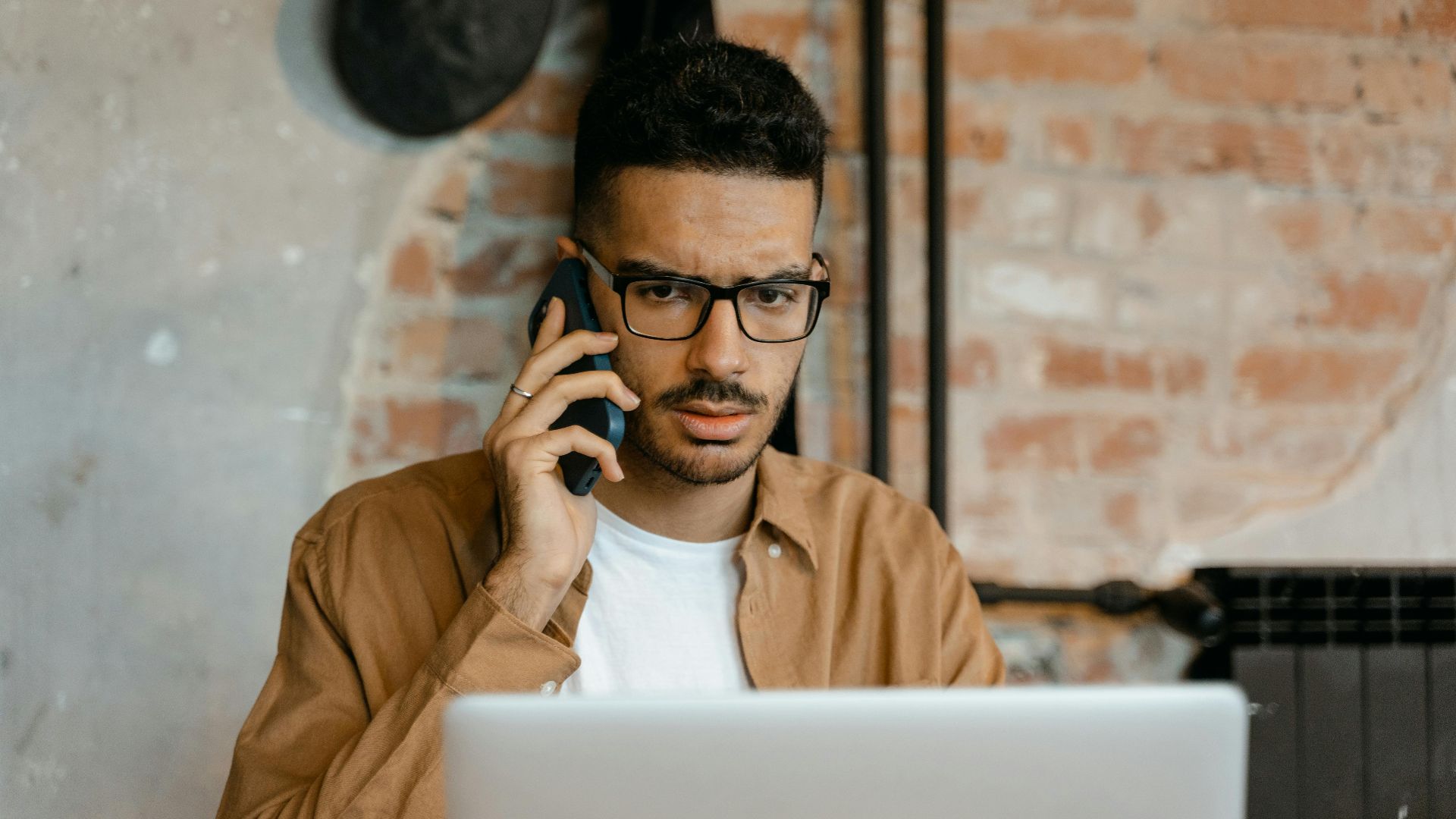Person with glasses on call using laptop, brick wall background.