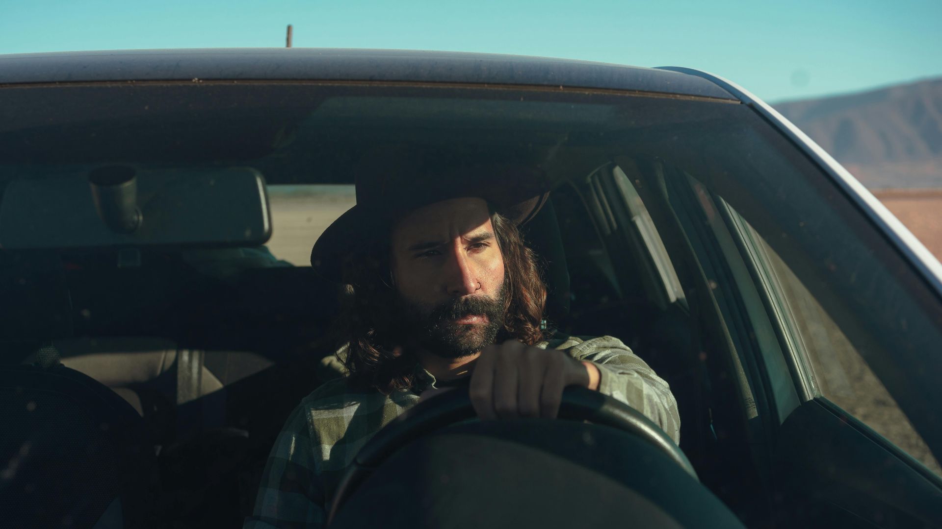 A bearded man in a hat driving a car through a sunny desert landscape, conveying a sense of adventure.