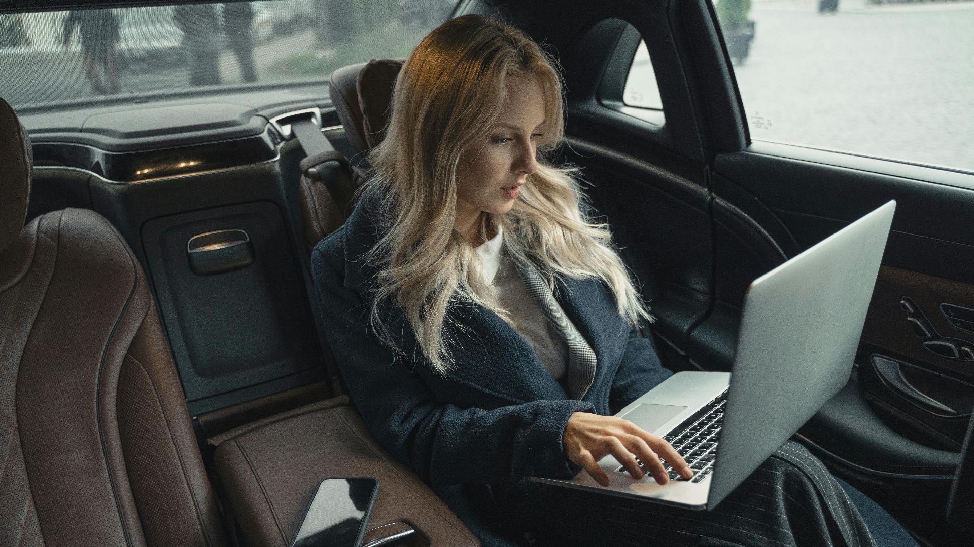 A woman in a luxury car's backseat working on a laptop with a smartphone nearby.
