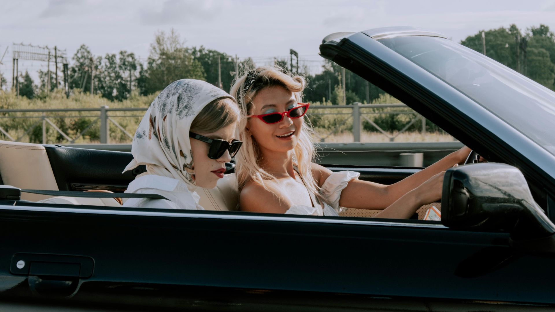 Two stylish women driving a convertible car outdoors, enjoying the sunny day with fashionable sunglasses.