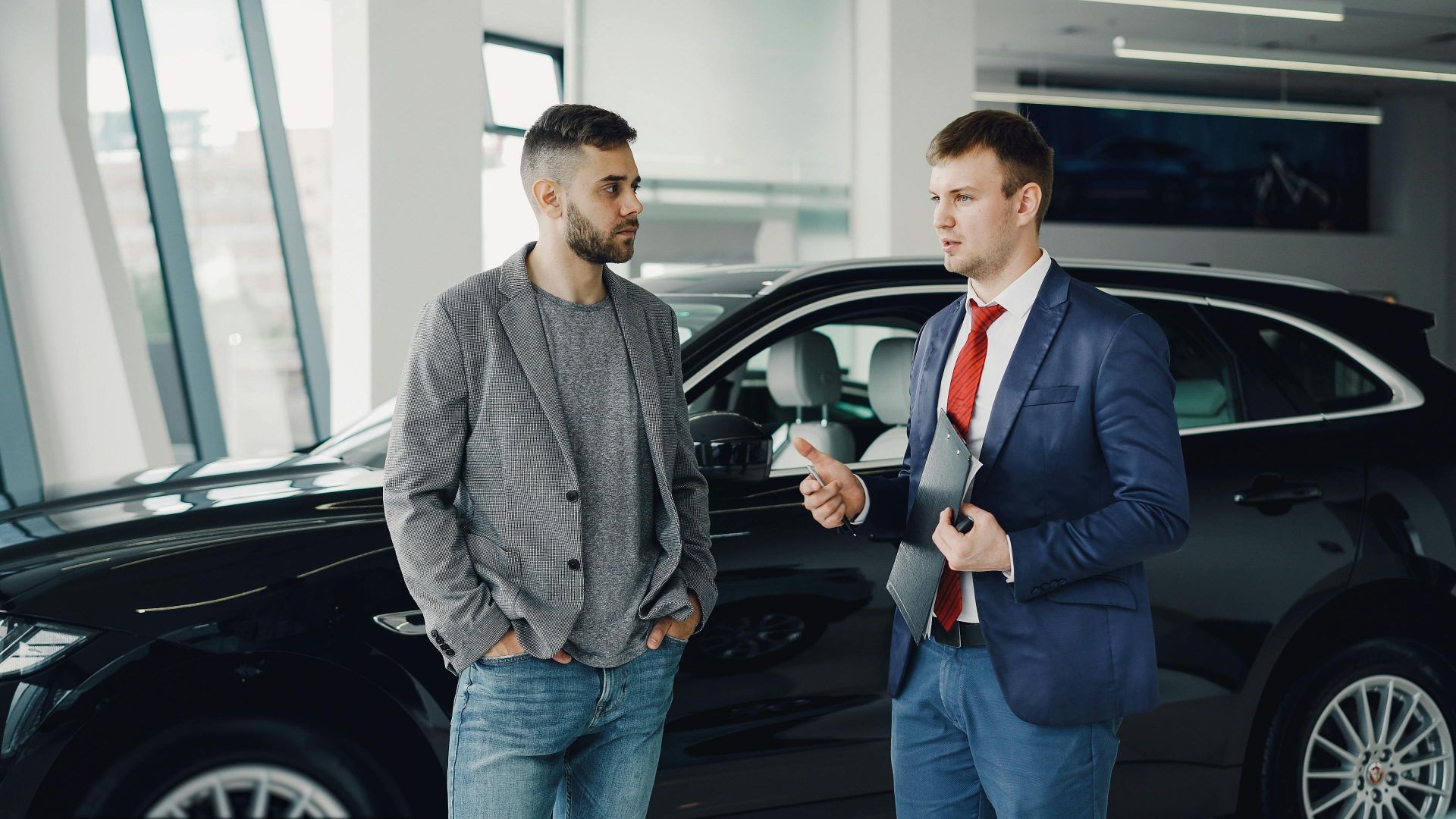 Two men discussing business in a car dealership, standing near a vehicle.