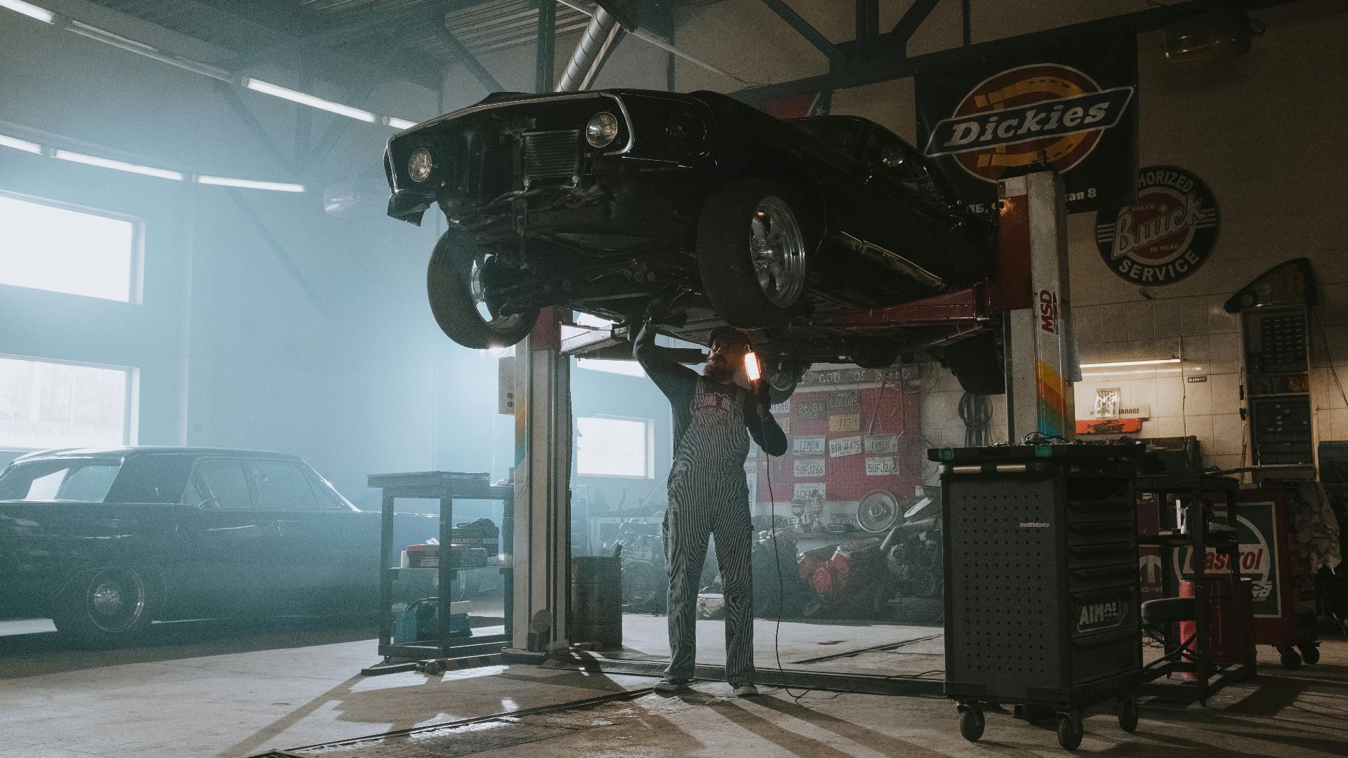Male mechanic using a light to inspect a classic car lifted in an automotive garage.