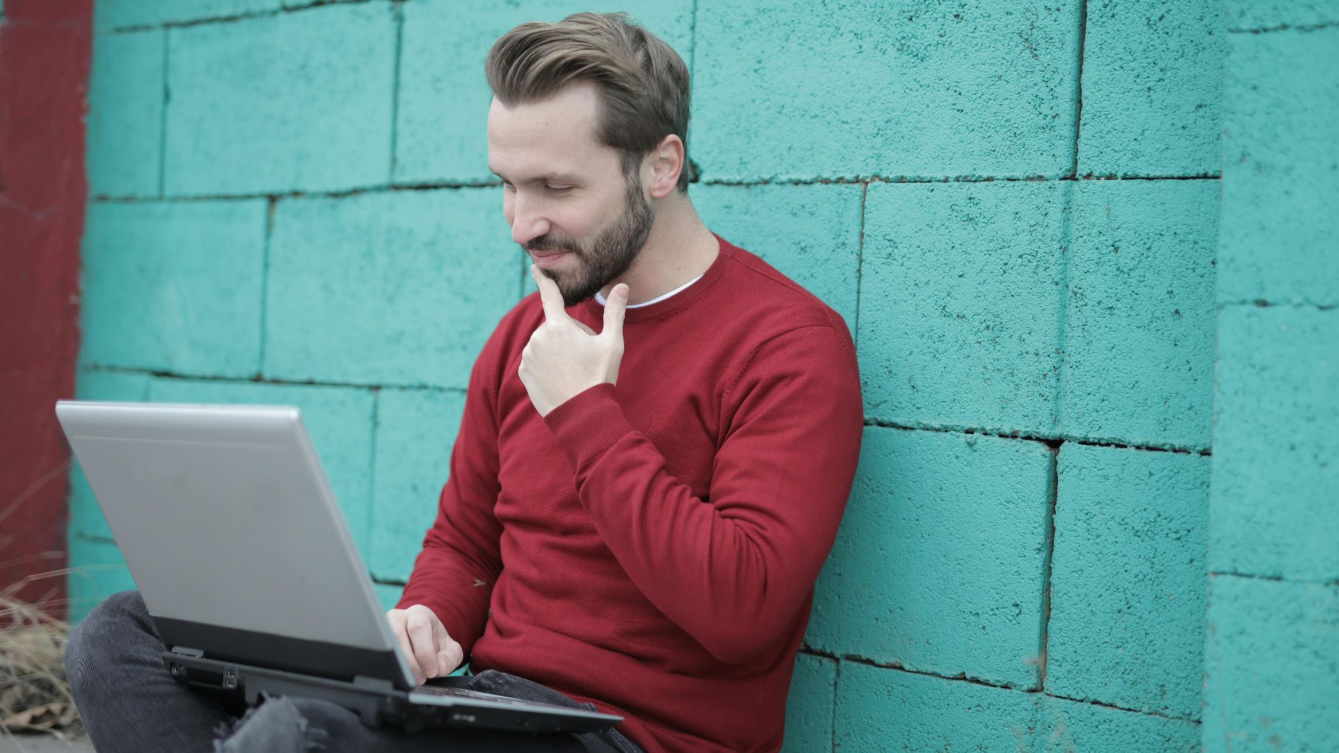 Smiling man using laptop for remote work, leaning on a vibrant green wall outdoors.