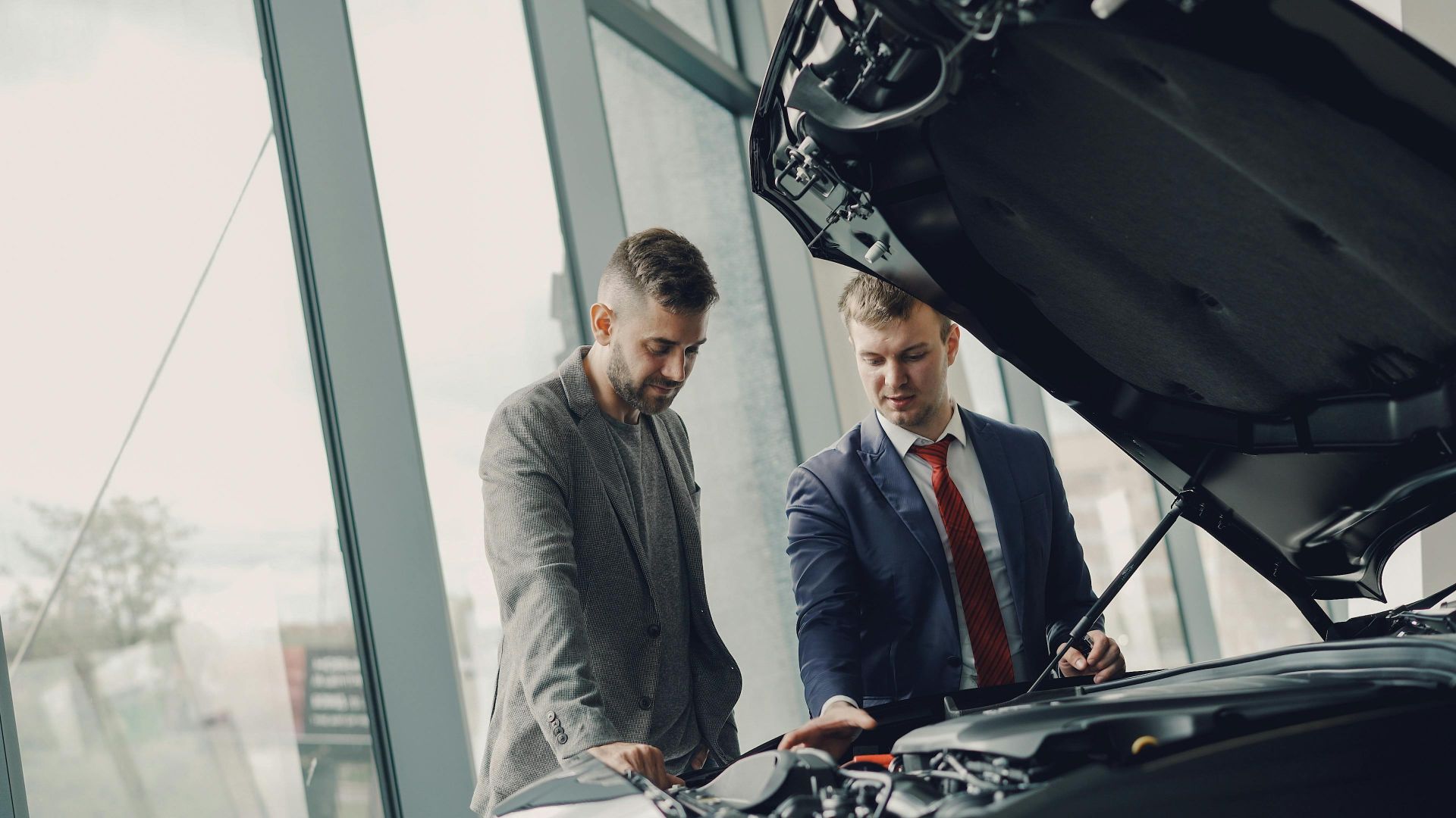 Two men inspecting a car's engine hood in a dealership showroom.