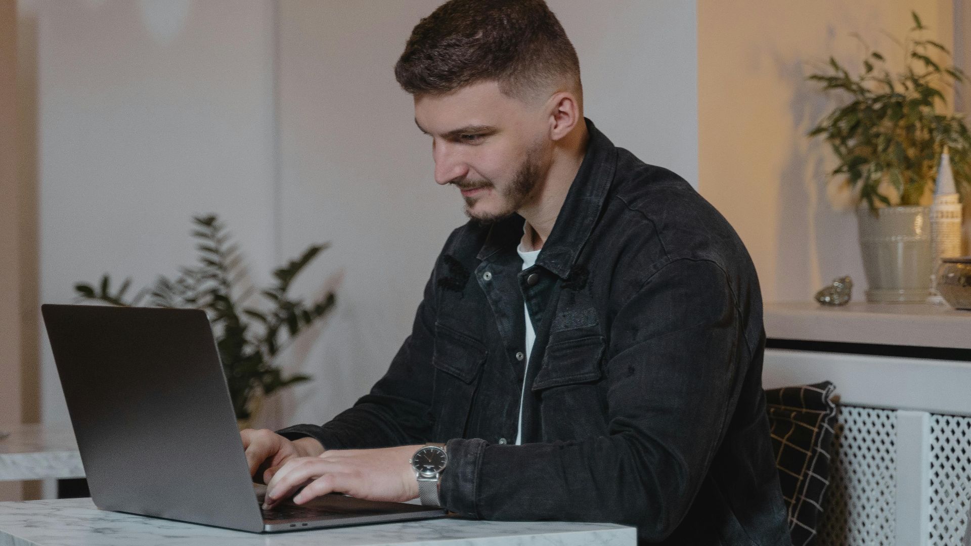 Caucasian man working on a laptop at a cozy indoor setting, surrounded by plants and candles.