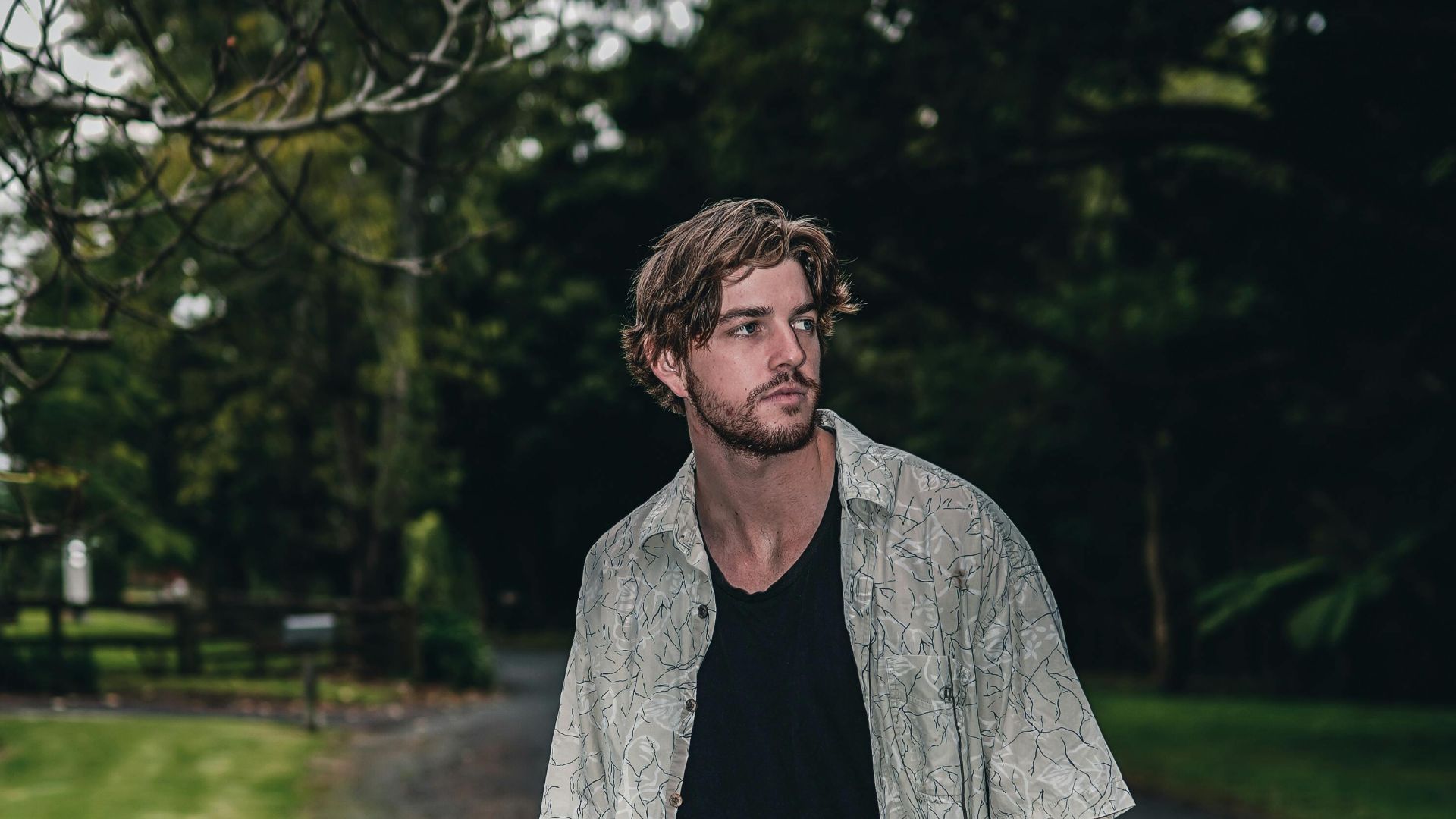 A trendy man in casual attire stands confidently on a forest pathway, embodying modern style.