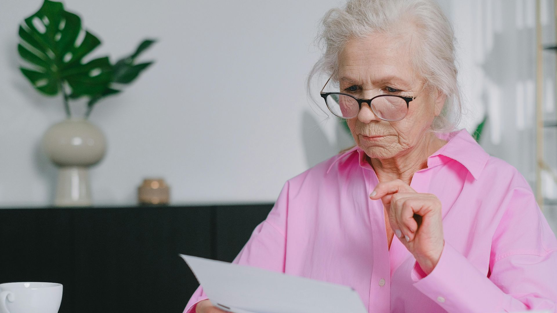 Elderly woman in pink shirt reading documents at home workspace.