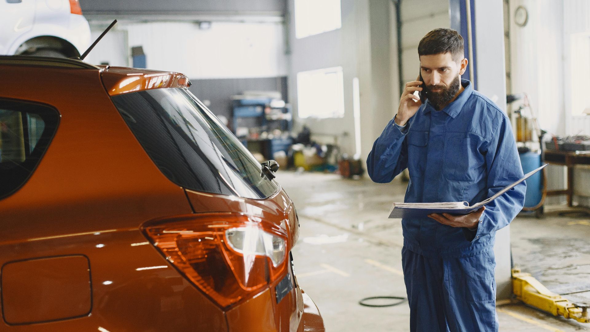 Mechanic in a blue uniform talks on phone beside a red car in an auto repair shop.